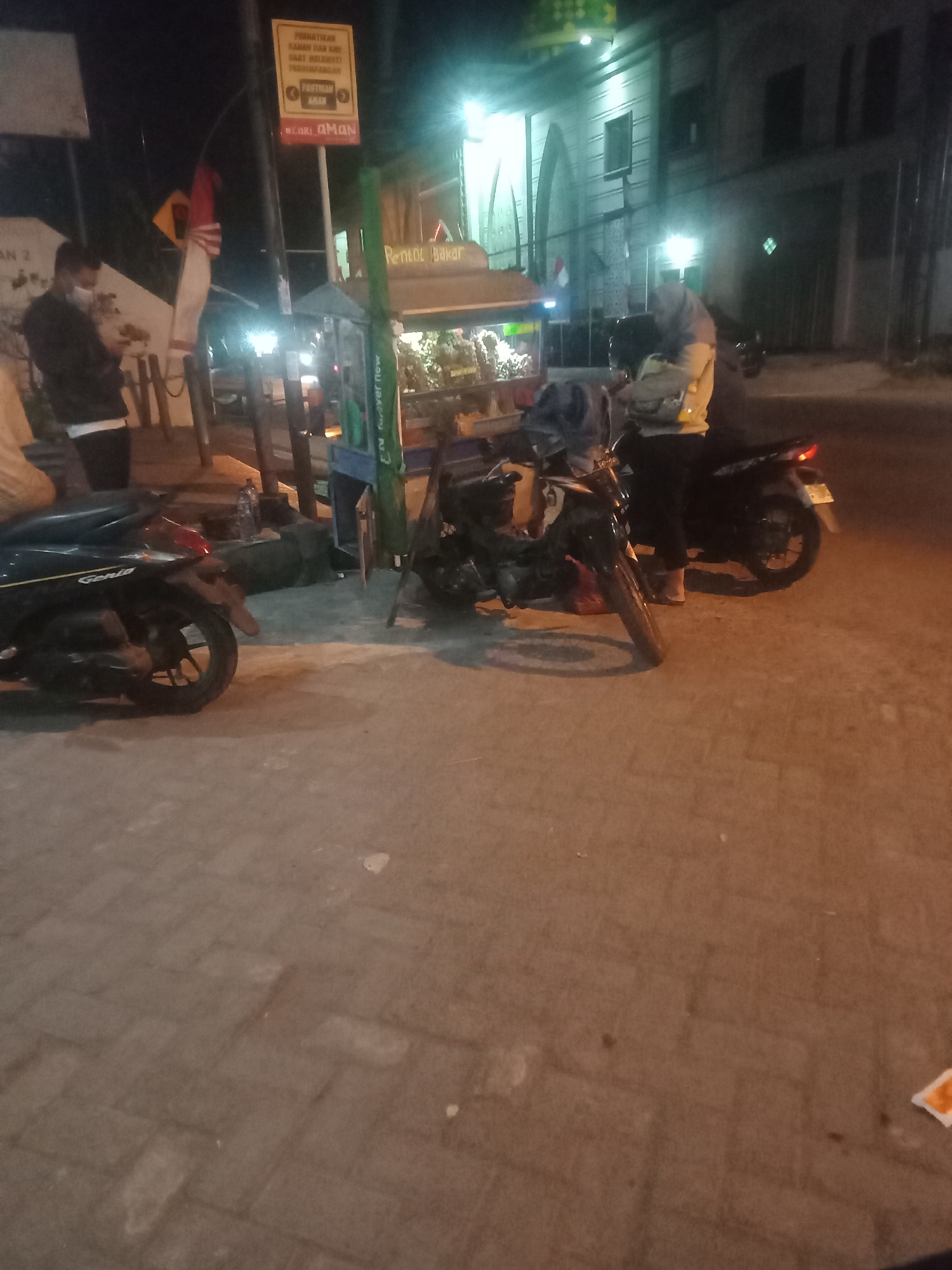 At night on a paved street in Katrungan, Indonesia, two motorcycles are parked. The motorcycle in the foreground is black and on the left side of the frame, with its rear wheel visible. The second motorcycle, also black, is to the right and slightly behind the first. A person wearing a grey hoodie and black pants stands next to the second motorcycle. In the background, a street vendor's cart with a green canopy is visible, with the text "Pantul Bakar" appearing on it. To the left of the cart, a person in a dark jacket and face mask is seen looking down at their phone. A street sign with a yellow warning symbol and red and white Indonesian flag is mounted on a pole. The street is illuminated by artificial lights, creating shadows on the paved surface.