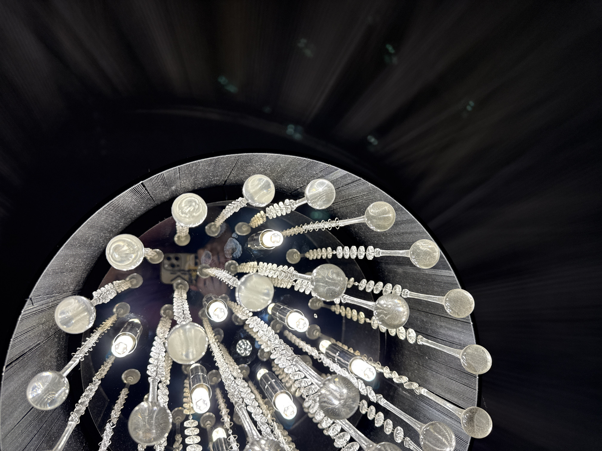 The image presents an upward-looking view of a modern chandelier, identified as being located in Berezyna, Ukraine. The chandelier features a circular, dark, and highly reflective central surface from which numerous clear glass or crystal elements descend. Among these elements are several small, illuminated cylindrical light bulbs, casting bright white light. Interspersed with the bulbs are decorative strands composed of small, textured, bead-like clear glass segments, each culminating in a larger, flattened, spherical clear glass piece.
A distinct reflection is visible on the central mirrored surface of the chandelier. This reflection clearly shows a person holding a smartphone, which is aimed upwards, indicating the individual is capturing the photograph. The reflection provides a partial view of the person's face, wearing what appears to be a light-colored top, and the rear camera array of the smartphone.
The upper perimeter of the chandelier's circular base displays a dark, finely ribbed or textured material. The background surrounding the chandelier is predominantly dark, almost black, with subtle, blurred streaks or light artifacts appearing on the right side of the frame, indicating a dimly lit environment beyond the chandelier's immediate vicinity.