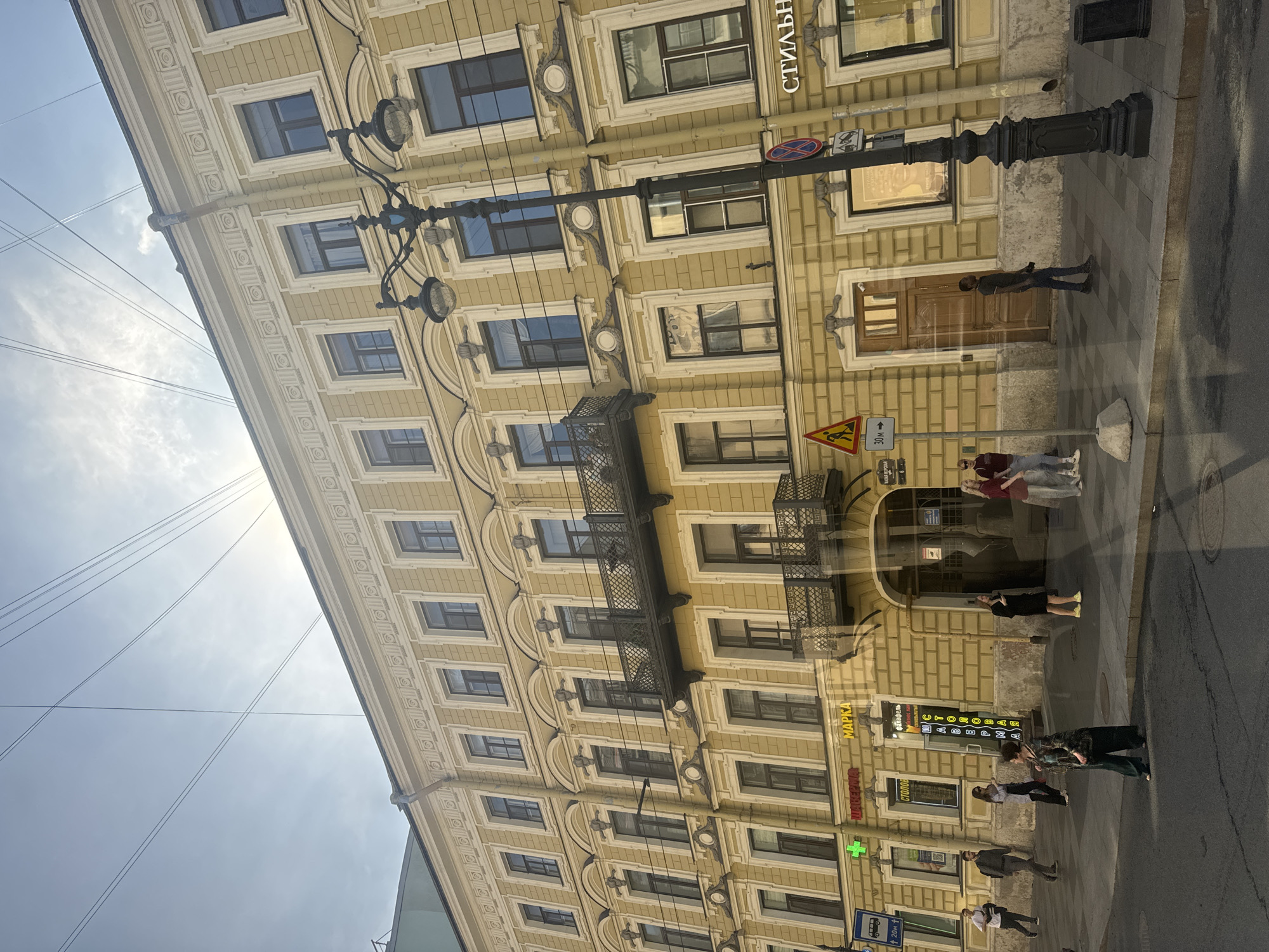 The image depicts a street scene in Saint Petersburg, Russia. A large, yellow, multi-story building with classical architectural elements dominates the frame. The building features numerous windows with dark frames, decorative moldings, and balconies with ornate railings. Several signs with Cyrillic script are visible on the building's facade, indicating businesses or shops. A lamppost stands on the sidewalk in front of the building. Several people are walking on the sidewalk and crossing the street. Overhead, power lines crisscross the sky, common in many urban environments. The sky is bright, suggesting daytime.