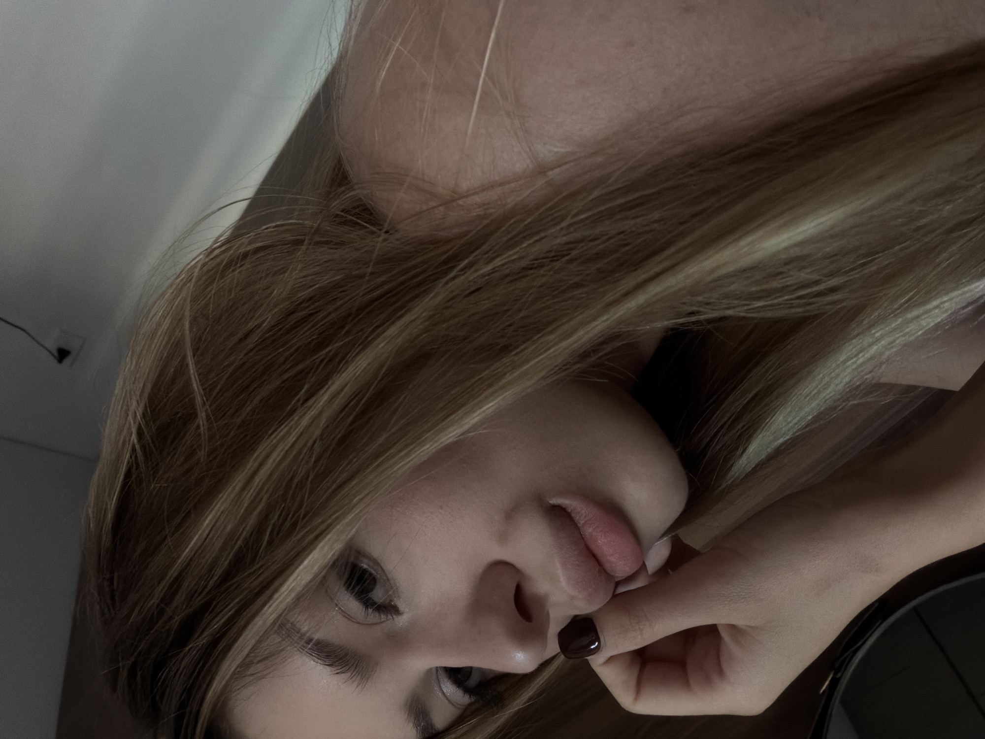 The image is a close-up, self-taken photograph of a young woman's face, with her head angled and her hand near her mouth. She has blonde hair with lighter streaks, fair skin, and is wearing dark nail polish. The background is indistinct, appearing to be an indoor setting with a light-colored wall and ceiling. There is no discernible information about the city of Da Nang, Vietnam, in this image.
