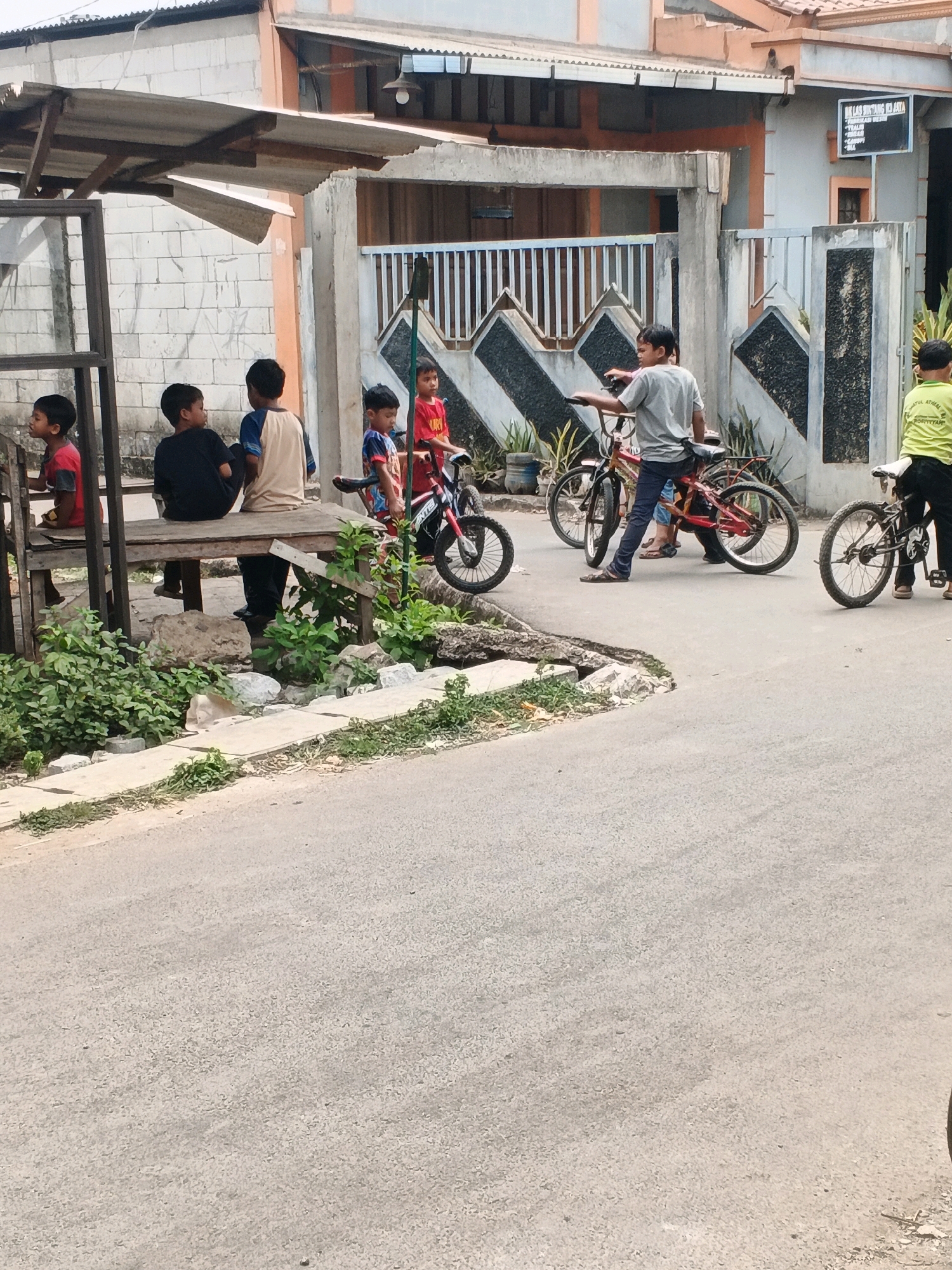 The image depicts a street scene in Kadu, Indonesia, featuring a group of children with bicycles near a simple outdoor shelter. In the foreground, a paved road is visible with a concrete drainage channel running along its edge, partially overgrown with green vegetation and scattered stones. To the left, a makeshift wooden shelter with a corrugated metal roof provides shade. Underneath the shelter, four young boys are seated on a wooden bench. One boy, wearing a red t-shirt, is seated on the far left. Next to him, another boy in a black t-shirt is looking away. A third boy, wearing a beige and blue t-shirt, sits in the middle, and a fourth boy in a black t-shirt is partially visible on the right.
Outside the shelter and further into the scene, several other children are interacting with bicycles. A boy in a red t-shirt and patterned shorts stands with a red bicycle. To his right, a boy in a grey t-shirt and dark pants stands with a red bicycle, appearing to gesture with his left hand. Another boy, wearing a light green t-shirt, stands to the far right with a bicycle. In the background, a building with light-colored walls and an orange trim is visible. A fence with vertical white bars and darker decorative elements separates the building from the street. A black sign with white text is mounted on the wall of the building. The overall environment appears to be a residential or semi-commercial area, characterized by simple structures and outdoor gathering spaces.