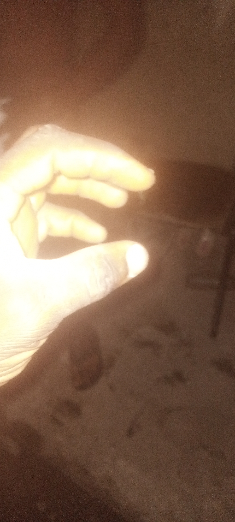 A hand is visible in the foreground, with outstretched fingers. The lighting is poor, but the hand appears to have darker skin. The background is out of focus, but a table with items on it and a chair leg can be discerned. There is no information present to confirm location or reference Sabon Tasha, Nigeria.