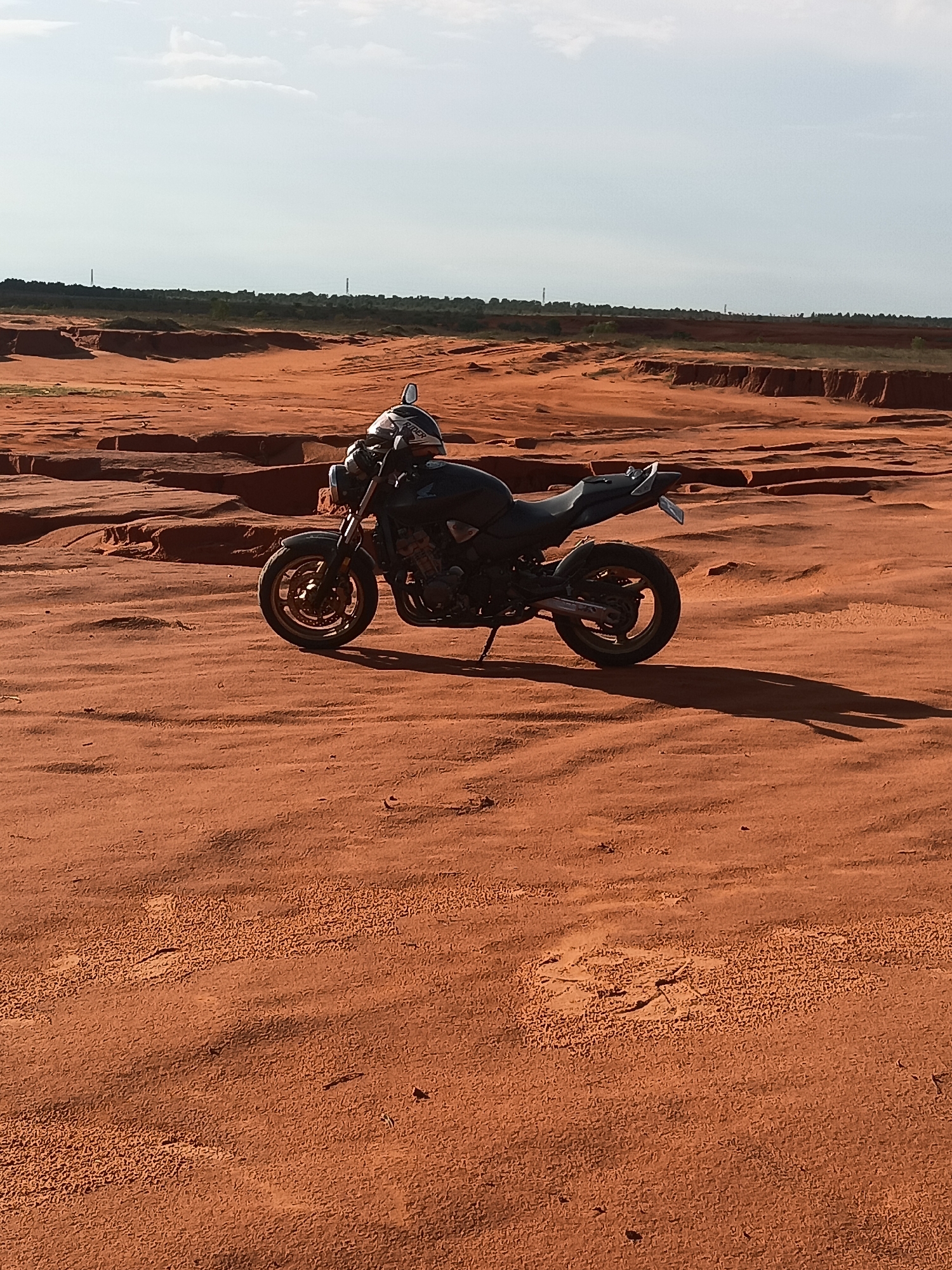 A black motorcycle with a silver and black helmet on its handlebars is parked on a sandy, red dirt landscape. The motorcycle is angled towards the left, with its front wheel on the left and its rear wheel on the right. The surrounding terrain is characterized by rolling sand dunes and layered red rock formations. In the distance, a line of trees and a hazy horizon are visible under a bright, overcast sky. The scene appears to be a natural landscape, possibly a desert or a dry, eroded area, and the presence of the motorcycle suggests it might be a recreational or adventurous location. The lighting suggests it is daytime, possibly late afternoon due to the long shadow cast by the motorcycle.