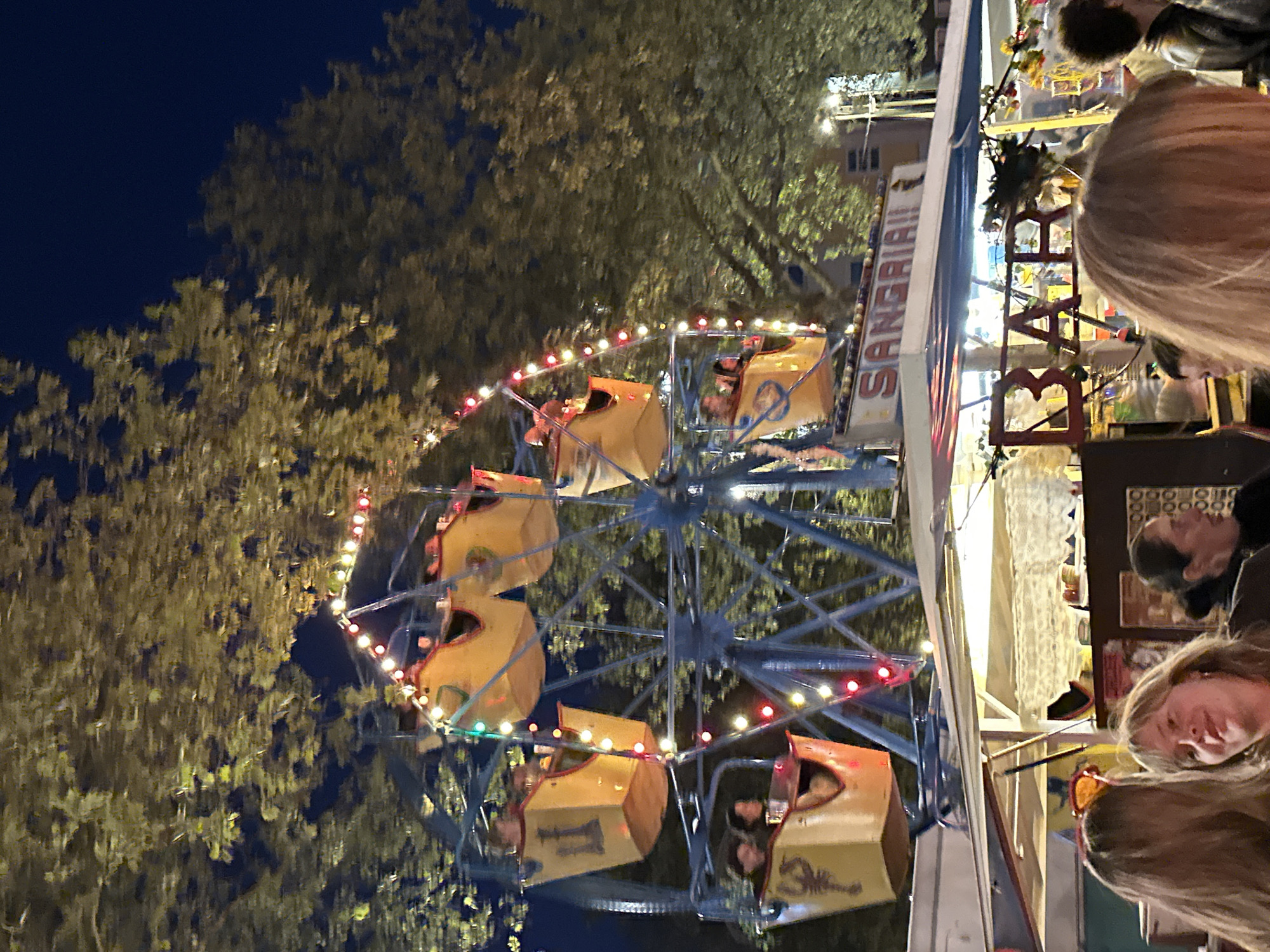 The image shows a ferris wheel at night with patrons riding in the cars. The ferris wheel is decorated with red, white, and green lights. In the foreground, there is a bar tent called "Sangria Bar." It is possible that this scene is taking place in Amsterdam, Netherlands.