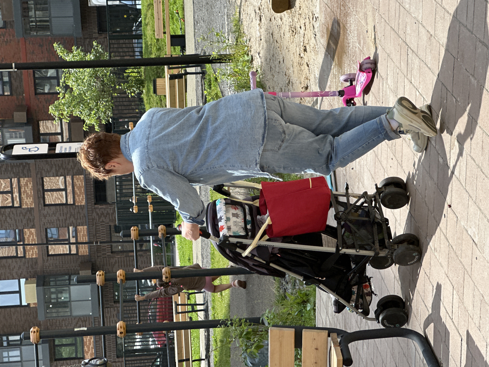 A woman with short reddish hair, wearing a denim shirt and jeans, stands in a playground. She is pushing a stroller with a red bag and a patterned fabric insert. A pink scooter is visible nearby. In the background, a child is hanging from a monkey bar on a playground structure. The playground is located in front of a modern brick apartment building. The ground is paved with light-colored bricks in the foreground, transitioning to sand in the playground area. The scene appears to be in the city of Znamya Oktyabrya, Russia.
