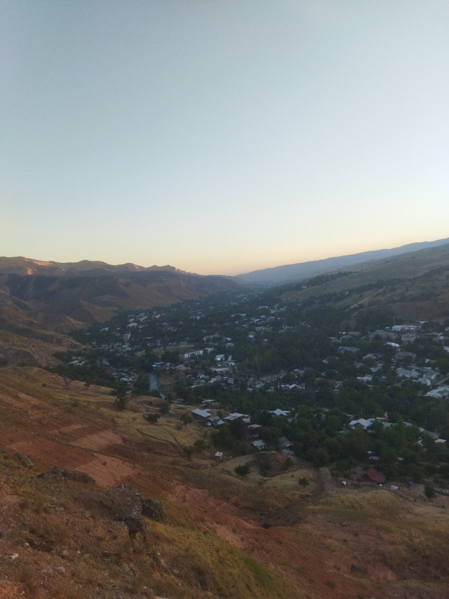 The image depicts a panoramic view of a valley in Mailuu-Suu City, Kyrgyzstan. In the foreground, a steep, rocky hillside covered in dry grass and reddish-brown soil slopes downwards. The midground features a dispersed settlement of houses with various roof colors, nestled among numerous trees. A winding river or stream is visible passing through the settlement. In the background, a series of rugged, barren mountains rise, with the hazy silhouettes of more distant ranges forming the horizon. The sky above is a pale, clear gradient from a light blue near the horizon to a lighter tone at the top of the frame, suggesting either early morning or late afternoon light. There are no visible people in the image. The scene is one of a rural or semi-rural town situated within a mountainous, arid landscape.