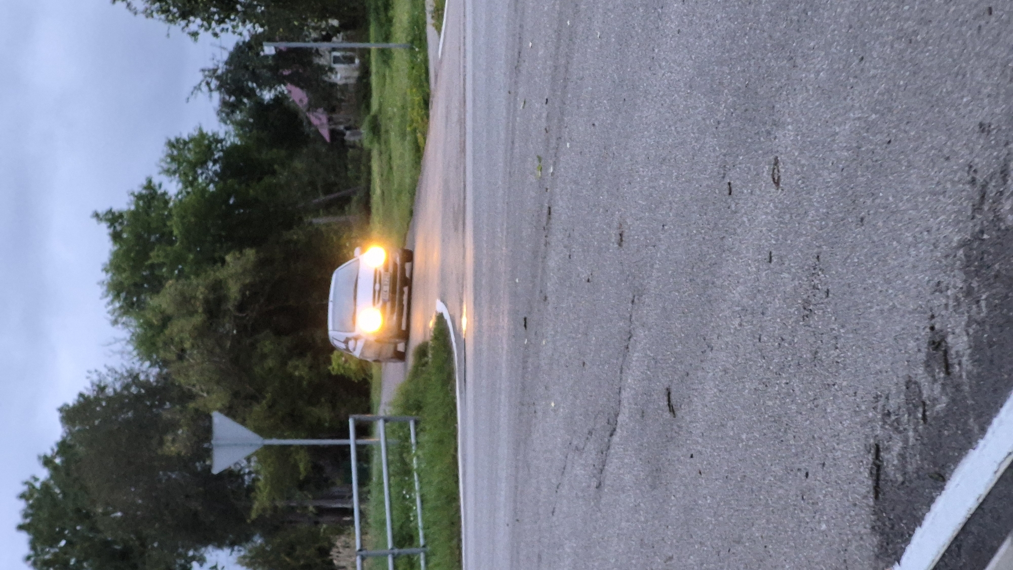 A silver hatchback car with its headlights on drives towards the viewer on a paved road. The car is positioned in the center of the frame and is approaching an intersection. Trees and vegetation are visible in the background, with a house and a signpost on the left side of the road. The sky is overcast, suggesting it is either dawn or dusk. The foreground shows a section of the paved road with some debris. The location is identified as Krauja, Latvia.