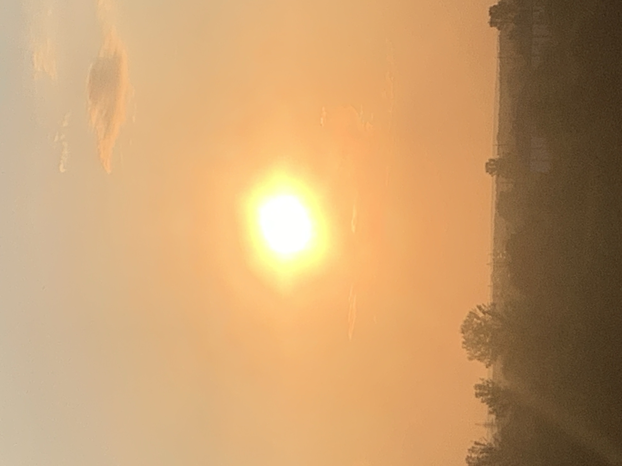 The image shows a sunset over Kolomyia, Ukraine. The sun is a bright, glowing orb in the center of the frame, casting a warm, orange light across the sky. There are a few wispy clouds scattered in the sky, one particularly noticeable cloud is in the upper right corner. Below the sky, a silhouette of trees and buildings can be seen against the vibrant sunset. The overall scene depicts a peaceful, natural landscape transitioning into dusk.
