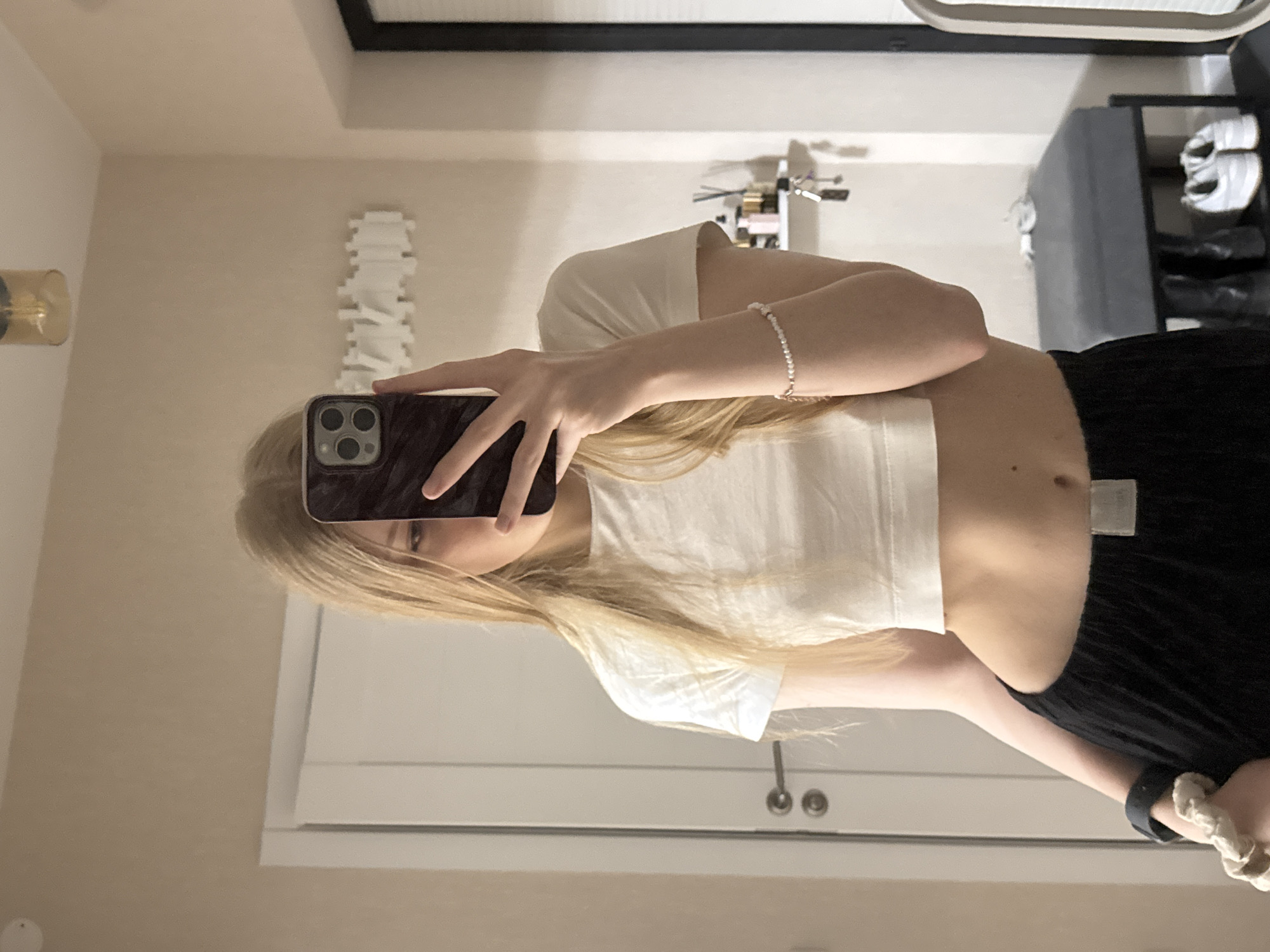 A young person, appearing female, with long, straight blonde hair, stands in an indoor setting, taking a mirror selfie. The image is taken from a slightly low angle, showing the person from the chest down, with the upper part of their face partially obscured by the smartphone they are holding.
The subject is wearing a white, short-sleeved crop top and dark-colored pants or a skirt. A delicate, light-colored beaded bracelet is visible on their right wrist, while their left wrist has a dark band and a white, fabric scrunchie. The smartphone, a dark model with a triple camera cluster on the back, is held up to capture the reflection.
The background, visible in the mirror, reveals a clean and modern room with light-colored walls, likely off-white or beige. On the left, a small, gold-toned light fixture or sconce is mounted on the wall. To the right of the subject's head, a distinctive white decorative item is affixed to the wall, appearing as a stack of interconnected, geometric or organic shapes, possibly resembling a stylized spine or an abstract sculpture. Further to the right, a dark shelf or bench holds several pairs of shoes, including a pair of white sneakers and other dark footwear. In the lower part of the reflection, a white door or cabinet with two round, metallic handles is visible. At the top of the frame, a dark, thin border suggests the edge of the mirror or a piece of wall art.
The setting appears to be a personal space like a bedroom or dressing area, indicative of a modern home in Perm, Russia, though no specific visual cues confirm the city. The activity is a casual self-portrait. No text is visible in the image. The lighting is artificial, giving no indication of the time of day.