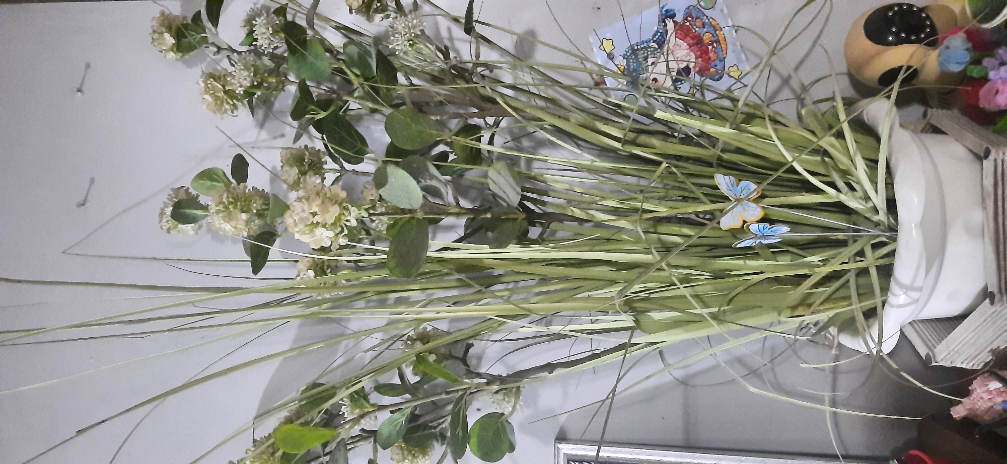 This is a medium-shot, eye-level, indoor photograph of a white ceramic planter filled with artificial greenery. The greenery consists of tall, thin, grass-like stalks interspersed with clusters of small, light-colored flowers and rounded, dark green leaves. Two decorative blue butterflies, one larger and one smaller, are perched on thin metal stems within the planter. To the right of the planter, partially visible, is a decorative ceramic tile depicting a cartoon-style figure. In the lower right corner, a portion of a tan, possibly stuffed, object with dark circular accents is visible. The background is a plain, light-colored wall with two small, metallic picture hooks. The overall scene appears to be decorative and artificial. The location is not discernible in this image and cannot be identified as being in Cisauk, Indonesia.