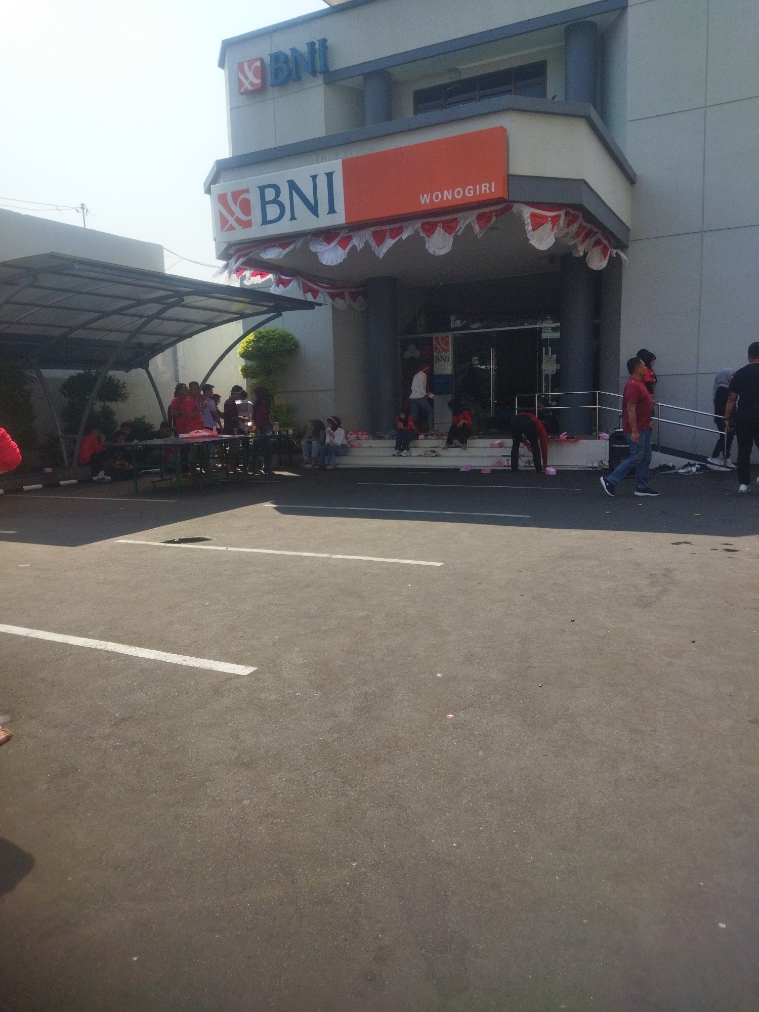 The image depicts the exterior of a BNI bank branch in Wonogiri, Indonesia. The building has a prominent orange sign that reads "BNI WONOGIRI." A festive red and white banner is draped across the entrance. In front of the bank, a group of people are gathered, some sitting on steps and others standing around tables. Several individuals appear to be interacting with each other or engaged in activities. The ground in front of the bank is paved with asphalt and marked with white lines. The overall scene suggests a public gathering or event taking place outside the bank.