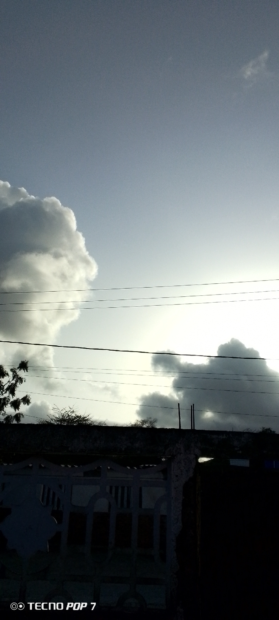 The image shows a cloudy sky with power lines stretching across the middle ground. Below the power lines, there is vegetation and a wall. In the foreground, there is a dark gate or fence. The image was taken in Altagracia, Venezuela. There are no people visible in the image.