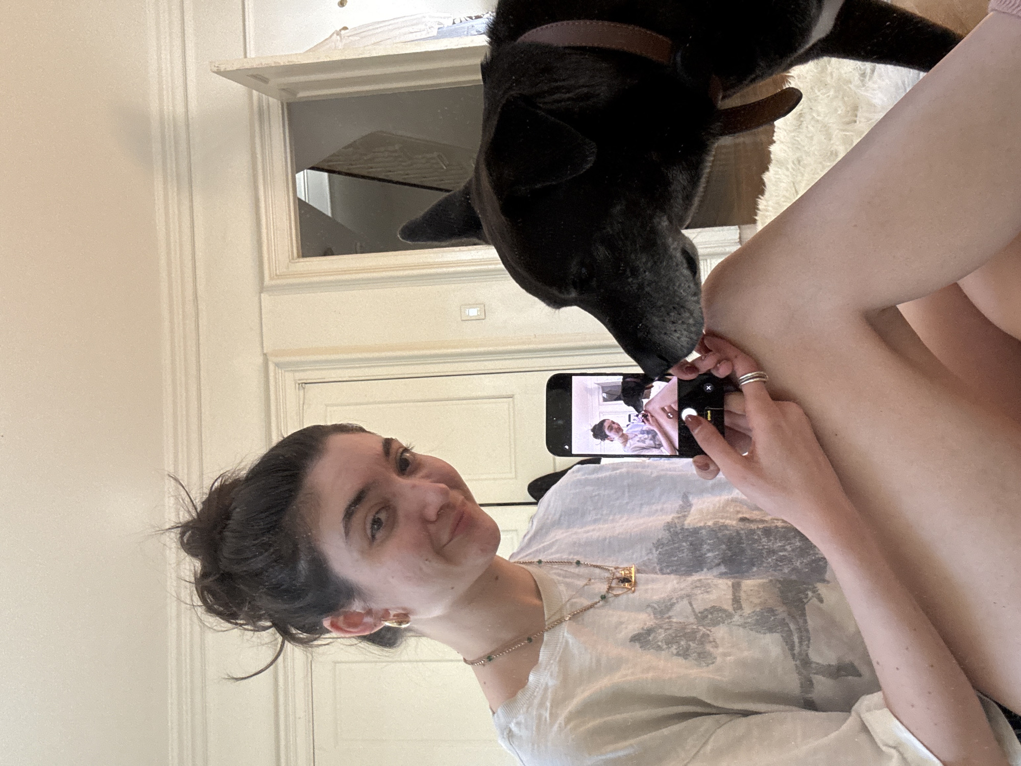 A young woman with dark hair tied in a bun is featured prominently in a bright, indoor setting, likely taking a selfie with her smartphone. She is smiling directly at the camera, wearing a light-colored t-shirt with a graphic design and a necklace. Several rings are visible on her left hand, which holds the phone.
A black dog with a brown collar is positioned above and to the right of the woman, its head lowered towards her hand or the smartphone, appearing to lick or nuzzle. The dog's interaction with the woman suggests an affectionate moment.
The smartphone screen shows the active camera interface, with options like "PHOTO" and "VIDEO" visible, along with "Live" and "2x" icons, confirming she is using the front-facing camera. The reflection on the phone screen shows a partial view of the woman herself.
The background consists of light-colored walls, white architectural trim, and what appears to be a transom window or opening above a doorway, indicating an interior space. A fluffy, light-colored rug is partially visible beneath the dog. The bright lighting suggests it is daytime. The overall mood is casual and warm, capturing a tender interaction between a person and their pet. The location context is New York, United States of America.