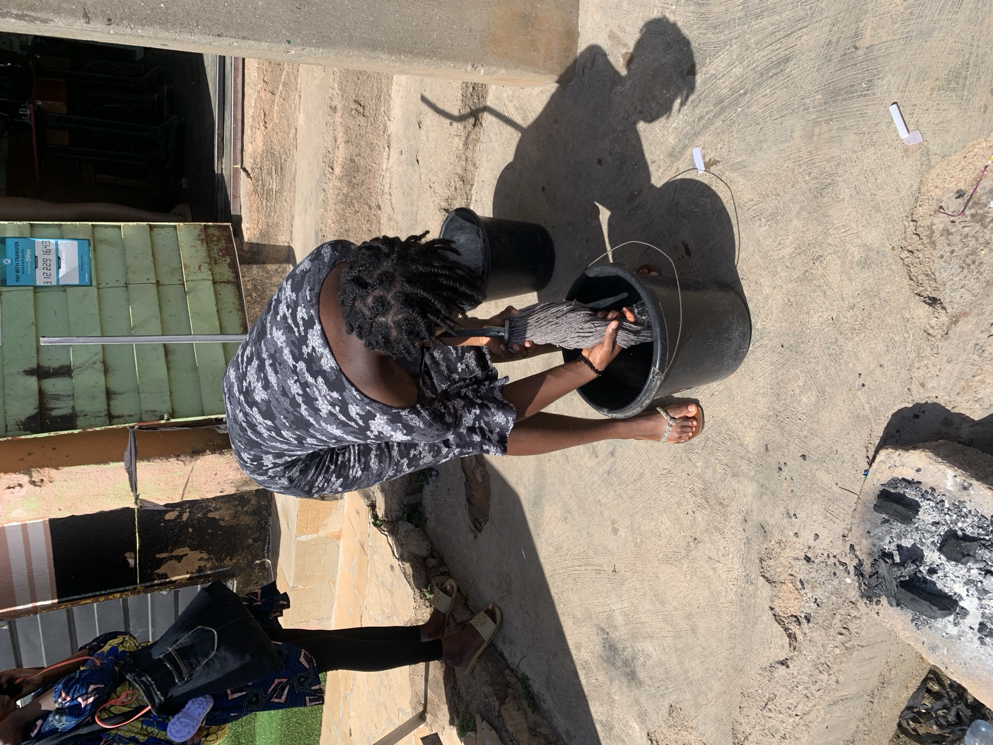 An overhead shot shows an adult female wringing out a mop into a black bucket. The woman has braided hair and wears a dark patterned dress, sandals, and a bracelet. Two black buckets are near her, one filled with a gray mop head. The scene takes place on a concrete surface, outside of a building. To the right of the woman and the buckets is an area that has a brick border and is filled with ash and charcoal. The location appears to be in Jos, Nigeria.