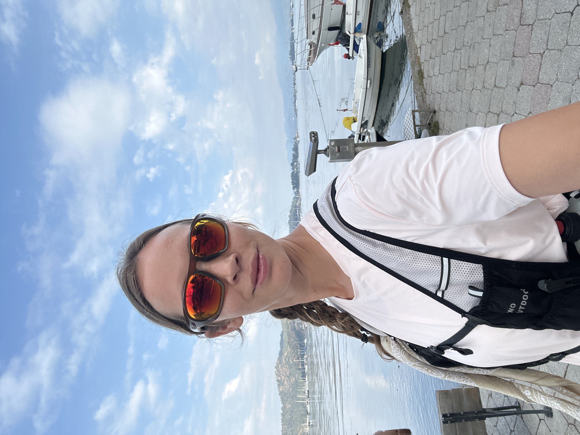 A woman is standing in Fethiye, Turkey, near a harbor with boats. She wears sunglasses, a light pink shirt, and a black hydration pack with a white logo. Behind her is a view of the harbor with boats, and in the background, there are mountains and a sky with scattered clouds. The woman is on a paved area near the water's edge, and a coin-operated telescope is also visible.