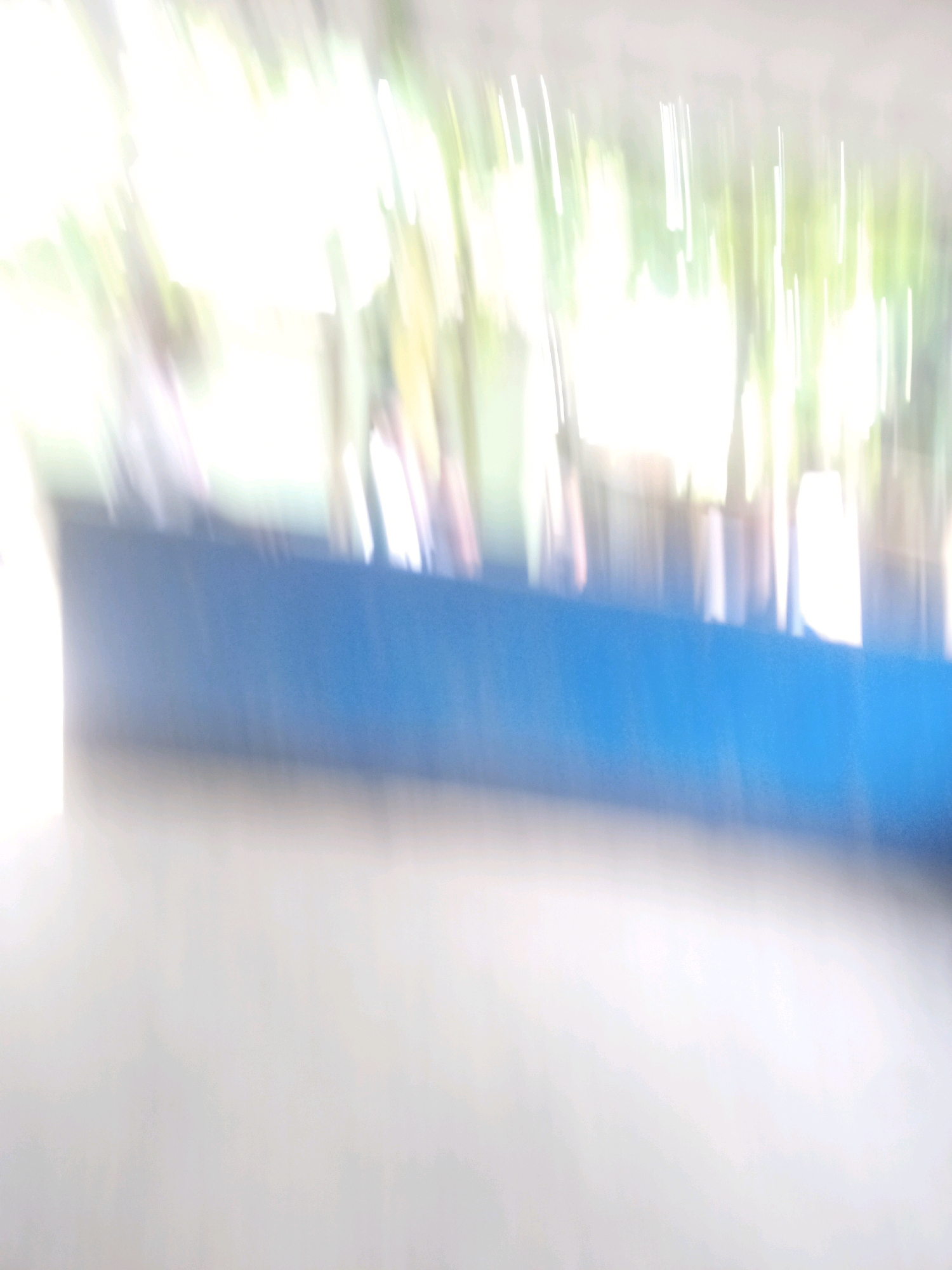 The image is a blurry, abstract depiction of an outdoor scene. A prominent horizontal band of bright blue dominates the mid-section of the image. Above this blue band, there are streaks of bright white and green, suggesting foliage or sunlight filtering through trees. Below the blue band, the image transitions to a lighter, blurred area that could be a surface or ground. The overall impression is one of motion blur, with vertical and diagonal streaks of light and color. There are no discernible people, subjects, or specific objects clearly identifiable due to the extreme blurriness of the photograph. The location in Hatsinigmari, India, is not visually evident from the image.