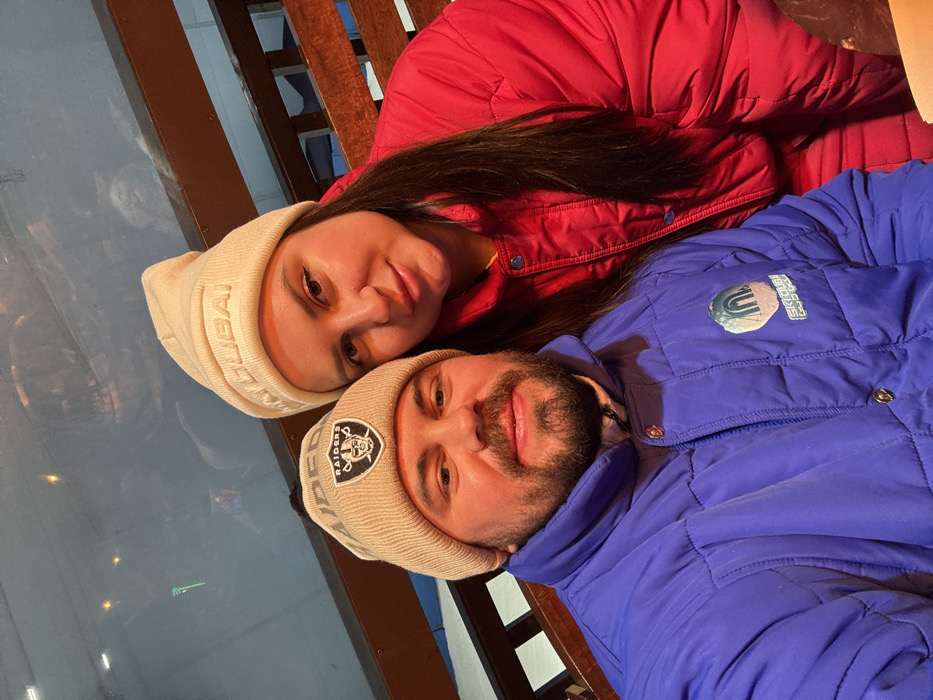 A male and female subject are depicted in a close-up, selfie-style photograph, looking directly at the camera. The male, positioned on the left, has a beard and is wearing a beige knit beanie with a black and white "Raiders" logo patch. He is also wearing a blue quilted puffer jacket, which features a "SKI DUBAI" logo patch on the left chest area. The female, positioned on the right, has long dark hair and is wearing a beige knit beanie with "SKI DUBAI" embossed text on the front. She is wearing a red quilted puffer jacket. The background behind the subjects consists of dark wooden slat structures, possibly benches or railings, and a reflective surface, likely glass, showing indistinct reflections. The specific location is Ski Dubai, an indoor ski resort situated within the Mall of the Emirates in Dubai, United Arab Emirates, as indicated by the "SKI DUBAI" branding on their clothing.