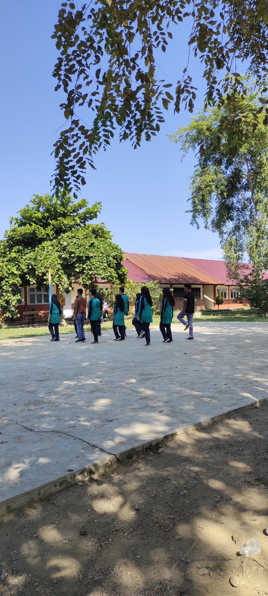 In Langnibong, Indonesia, a group of seven young people, four females and three males, stand in a line on a paved outdoor area. The individuals are dressed in casual clothing, with most of the females wearing turquoise tops and dark pants or hijabs. The males are dressed in a mix of casual attire, including a t-shirt and jeans, and turquoise tops similar to the females. They appear to be facing forward, possibly engaged in a group activity or waiting for instructions.
Behind them, a long, single-story building with a red-tiled roof is visible. The building has windows and doors, and appears to be a school or educational facility. Lush green trees are scattered around the area, providing shade. The ground is a mix of paved surfaces and dirt, with some scattered gravel. The sky above is clear and blue, indicating a sunny day. The overall scene suggests a relaxed outdoor environment, possibly during a school day or an organized event.