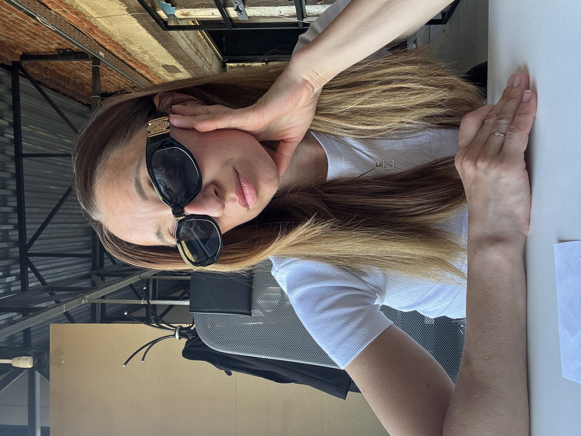 A woman is seated at a table, her right hand resting on its surface, with a ring visible on her ring finger. Her left hand is raised to her face, partially obscuring it, and she is wearing sunglasses. She has long, light brown hair. She is wearing a white shirt and a necklace. She is seated in an office chair. Behind her is a wall and ceiling with metal beams. The scene is located in the city of Chaikovsky, Russia.