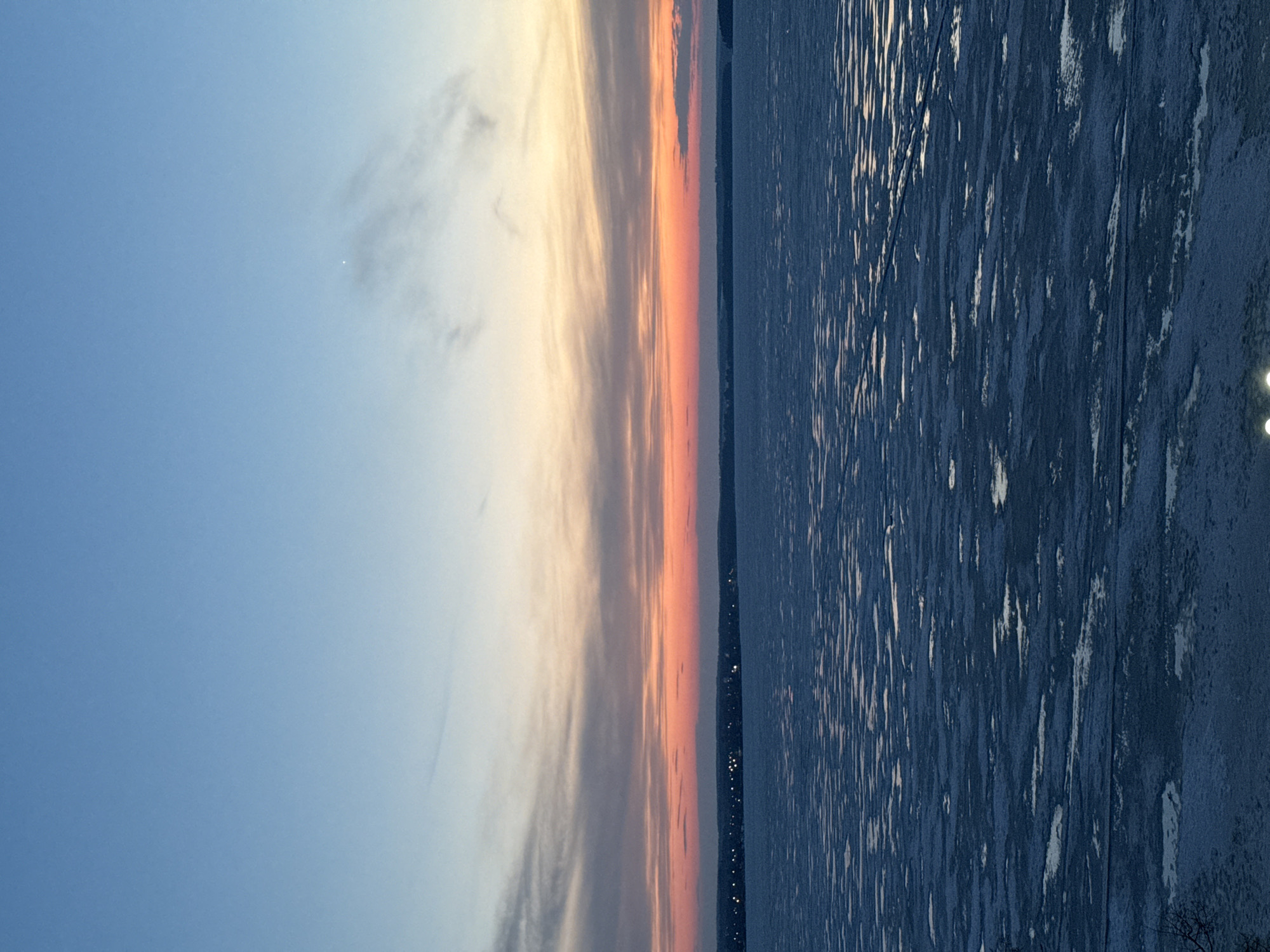 The image shows a sunset over what appears to be the Votkinsk Reservoir in Votkinsk, Russia. The sky transitions from blue to shades of orange and pink near the horizon. Clouds are visible in the sky. The body of water is dark, with light reflecting off its surface. A line of lights, likely from buildings, is visible along the horizon. The foreground contains a bank of land with a few scattered lights. The image is taken in a vertical format.