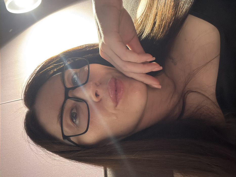 A close-up, rotated portrait of a woman looking directly at the camera. The image is rotated approximately 90 degrees clockwise, making it appear as if she is lying on her left side with her head towards the bottom of the frame and her body extending upwards.
The woman has long, dark brown hair that cascades around her. She is wearing black-framed glasses that sit on her nose, framing her eyes, which appear to be a light color. Her lips are slightly parted and have a shiny, perhaps glossed, appearance, with a light pink or nude color. There's a subtle highlight or reflection visible on her lower lip.
Her right hand is raised to the side of her face, with her fingers gently touching her cheek and jawline. Her nails are manicured with a natural, light polish. She appears to be wearing a dark-colored garment, possibly a top or dress, visible on her right shoulder and upper chest.
The lighting is bright and striking, with a prominent light source (possibly a lamp or window) visible in the upper left corner of the original image (or the top when upright). This strong light creates a bright halo effect and illuminates her face from above and slightly to the left, casting some shadows and highlights. The background behind her is light, possibly a wall, with the brightest area around the light source. The overall impression is that of a close-up selfie taken indoors under direct lighting. No text is visible in the image.