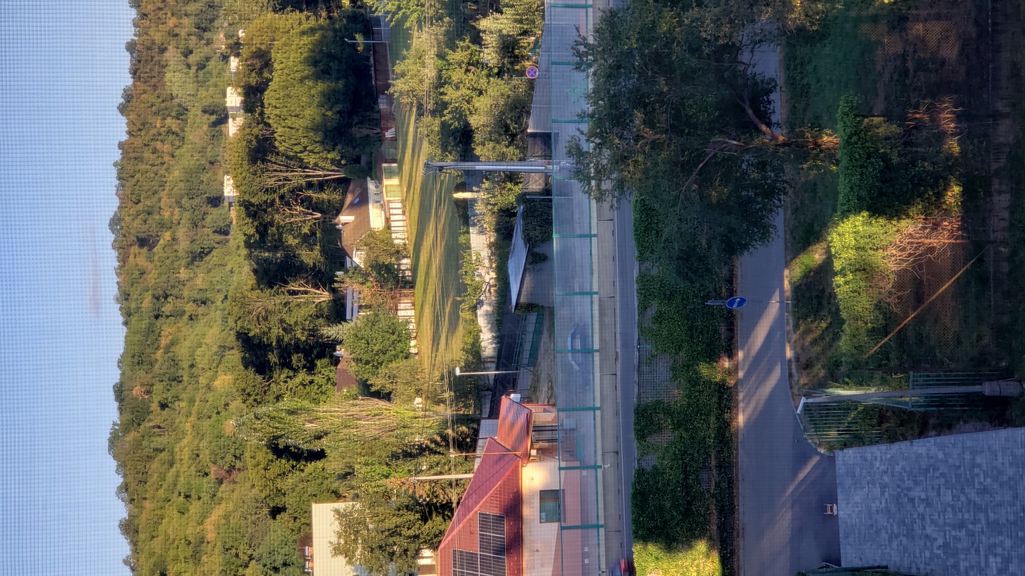 A wide-angle, high-angle outdoor shot shows a residential area in Prešov, Slovakia, with a lush, tree-covered hillside in the background. The foreground features a paved walkway leading down from the viewpoint, adjacent to a chain-link fence. A two-lane road runs horizontally across the mid-ground, separated from the houses by a green mesh fence. A few cars are visible on the road. To the left, a house with a red, gabled roof and solar panels is partially visible, with a smaller attached structure with an orange roof. Further back, several other houses with pitched roofs are scattered across a grassy lawn, bordered by fences and trees. The hillside is densely populated with trees, some showing the yellow and orange hues of early autumn. The sky above is a clear, light blue with a slight haze. The lighting suggests it is late afternoon, with long shadows cast by the trees across the road and grassy areas. A single blue circular traffic sign with a white arrow indicating "turn right only" is positioned on the side of the road.