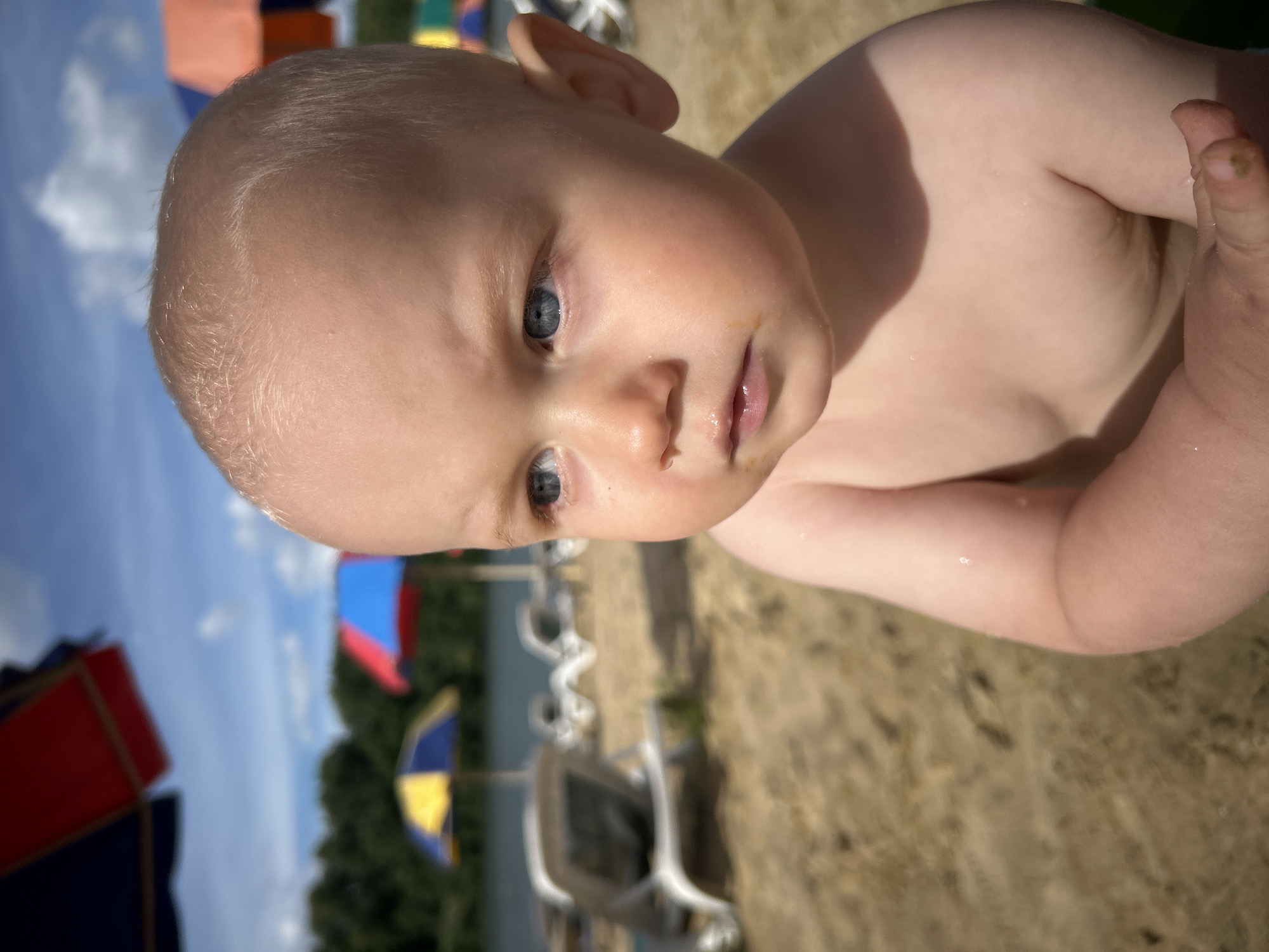 The media file shows a close-up, tilted shot of a baby's face and upper body. The baby has fair skin and light blonde hair. Their eyes are open and looking towards the viewer, and their lips are slightly parted. The baby's skin appears wet in places, and there is a small smudge of dirt on their finger. The background suggests an outdoor setting, with sand visible in the foreground and blurred elements of beach umbrellas and trees in the distance. The sky, also visible, appears blue with some clouds. The lighting indicates it is daytime, likely sunny. There is no visible text in the media file.