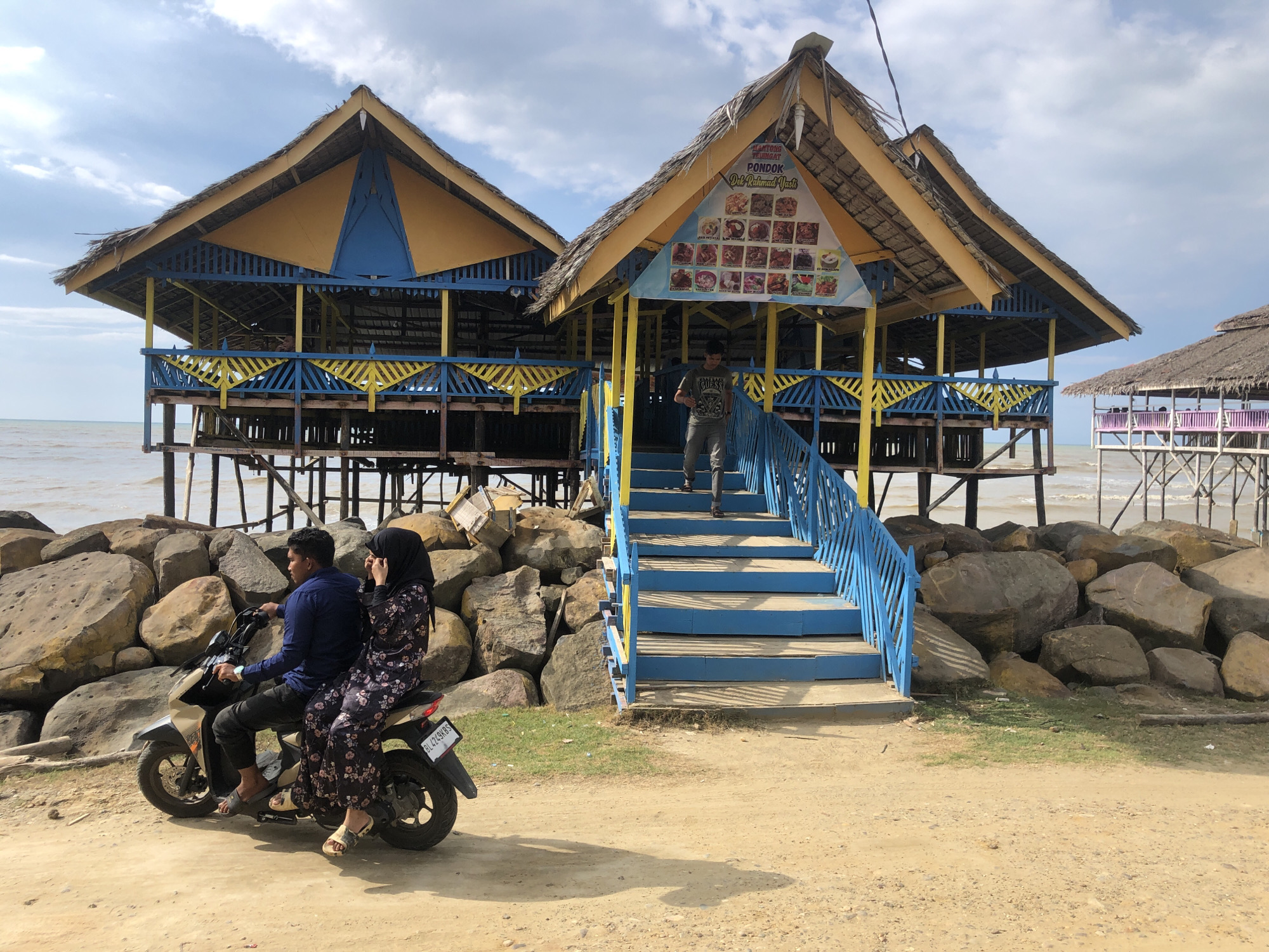 A sunny day at Lancok, Indonesia. Two stilt houses, painted yellow and blue, stand elevated above the shoreline rocks. A man is descending the blue steps of the right-hand structure, which is adorned with a menu. A couple is on a scooter facing away from the viewer in the foreground on a beach in front of these structures. The man wears a dark blue shirt and black pants, while the woman is in a floral dress and a black hijab. The license plate on the scooter is visible. In the distance, another similar stilt house is visible on the shoreline. The sky is partly cloudy.