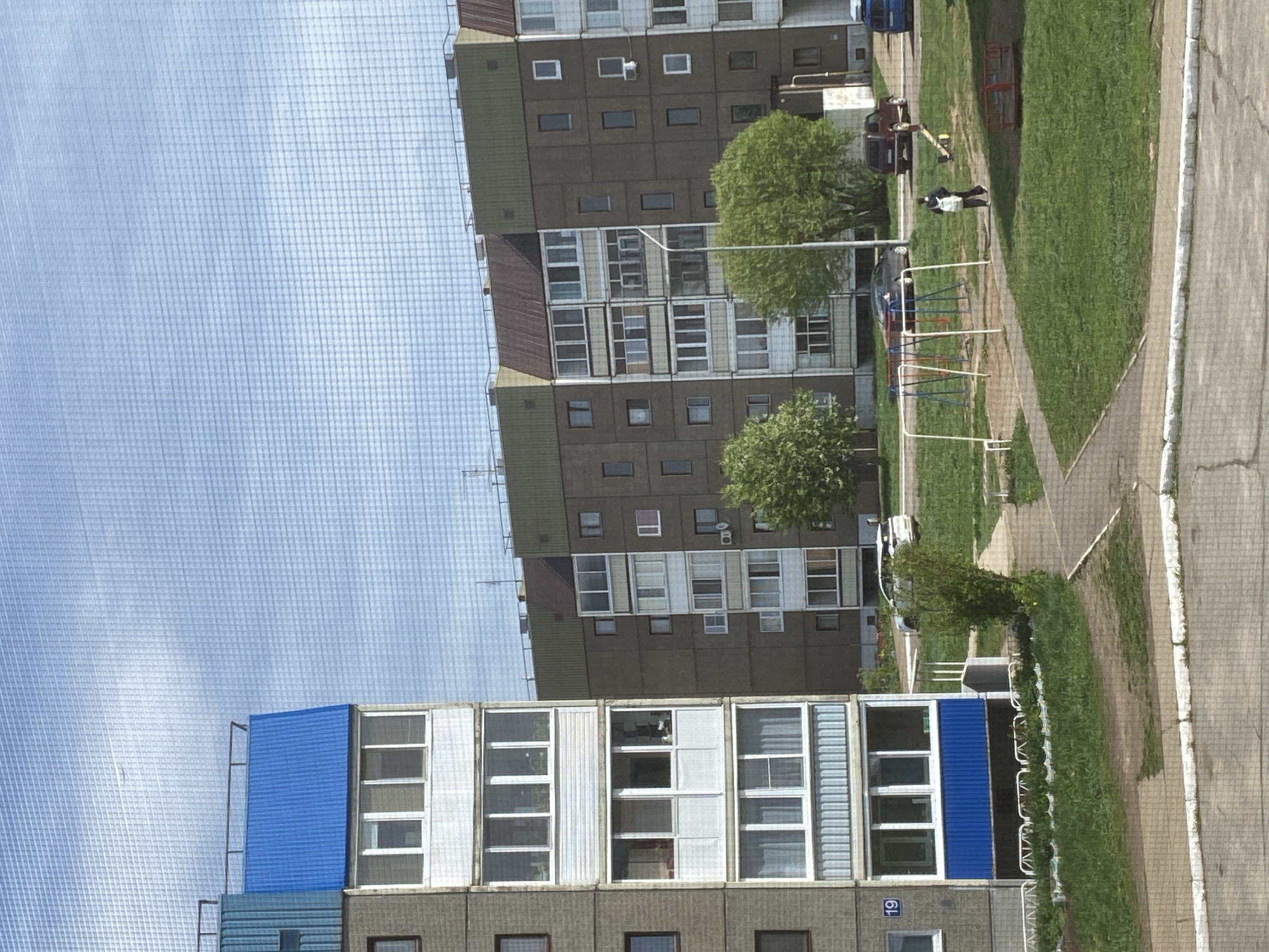 The image depicts a cityscape in Марковский, Russia. Two tall apartment buildings dominate the scene. One building is grey with a blue roof. The other is grey and brown. A chain-link fence covers the sky. Between the buildings is a green lawn and sidewalk. A person is walking along the sidewalk. A playground is visible in the background. Cars are parked near the buildings. Several trees are on the lawn.