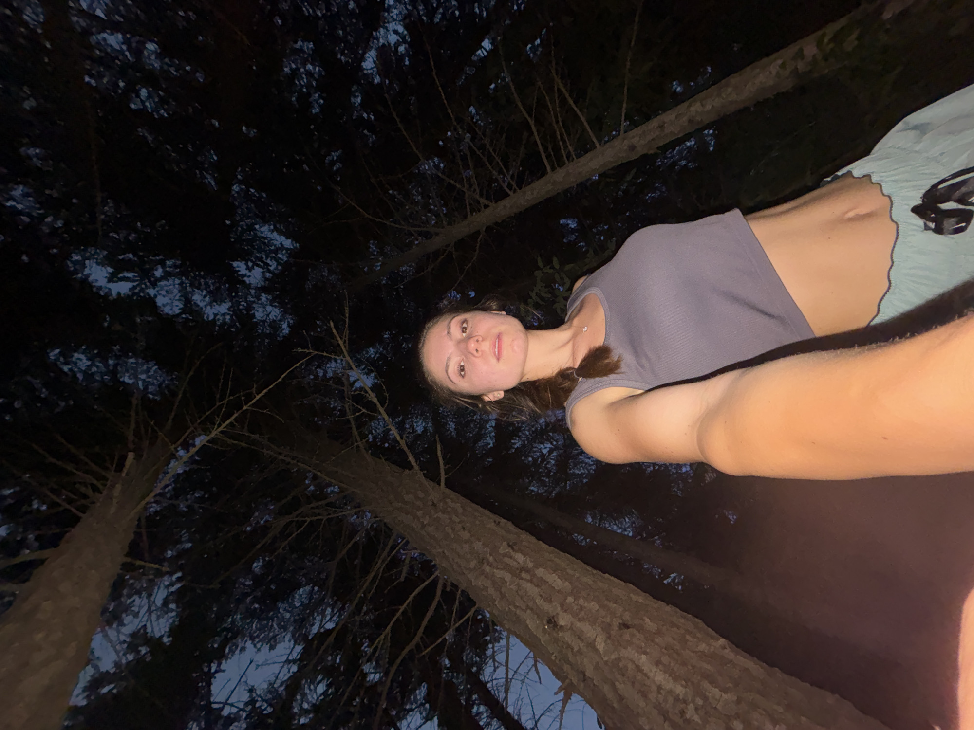 A young Caucasian female is taking a selfie outdoors at night. She is positioned in the lower right quadrant of the image, facing the camera. She has her left arm extended, holding the camera. She is wearing a cropped grey tank top and light blue shorts with a scalloped hem. Her hair is pulled back into a braid. She is standing amongst tall trees with dark foliage and a dark sky visible above. The image appears to be taken in a forest setting. There are no indications that this location is in the city of Varna, Bulgaria.