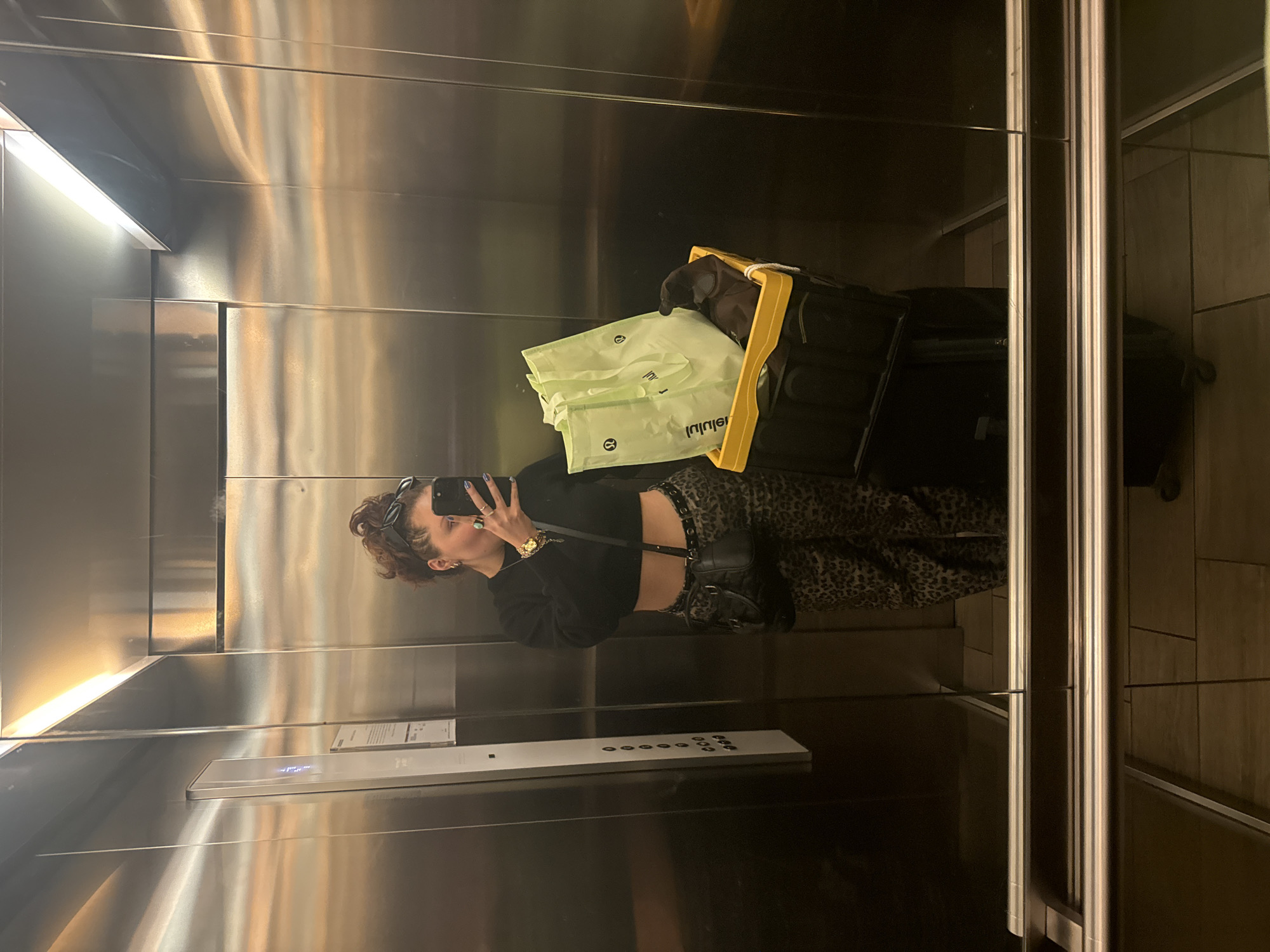 The image is taken inside an elevator, reflecting in the metallic walls. A woman wearing sunglasses, a black long-sleeve shirt, and patterned pants stands in the elevator, holding a phone up to take a selfie. Next to her is luggage. The luggage has bags from Lululemon. The elevator has a panel of buttons. Through the elevator doors, a tiled wall is visible. Based on the luggage, the woman appears to be a traveler. The precise location within Mexico City, Mexico cannot be determined from the image.