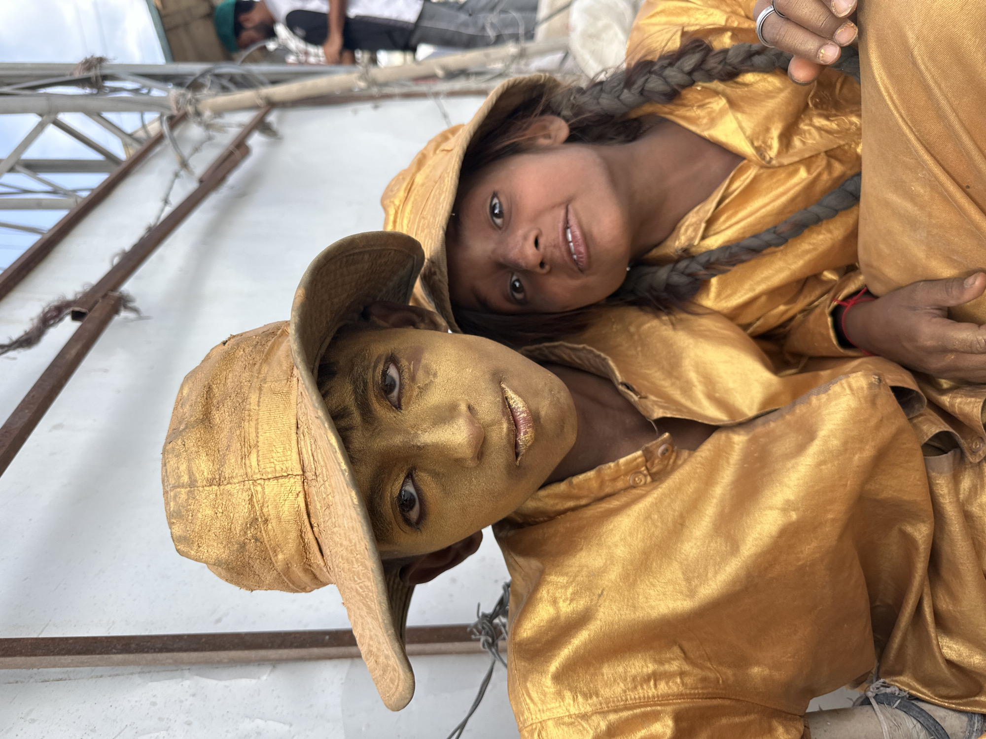 Two people are shown together in Quetta City Tehsil, Pakistan. The person in the foreground has gold-painted skin and wears a hat and a gold-colored jacket. The person in the background has unpainted skin and wears a gold-colored hooded jacket, and the person's hair is styled in a braid. They appear to be sitting, and the environment behind them includes metal framing, a white surface, and at least one person with a green hat in the upper portion of the image.