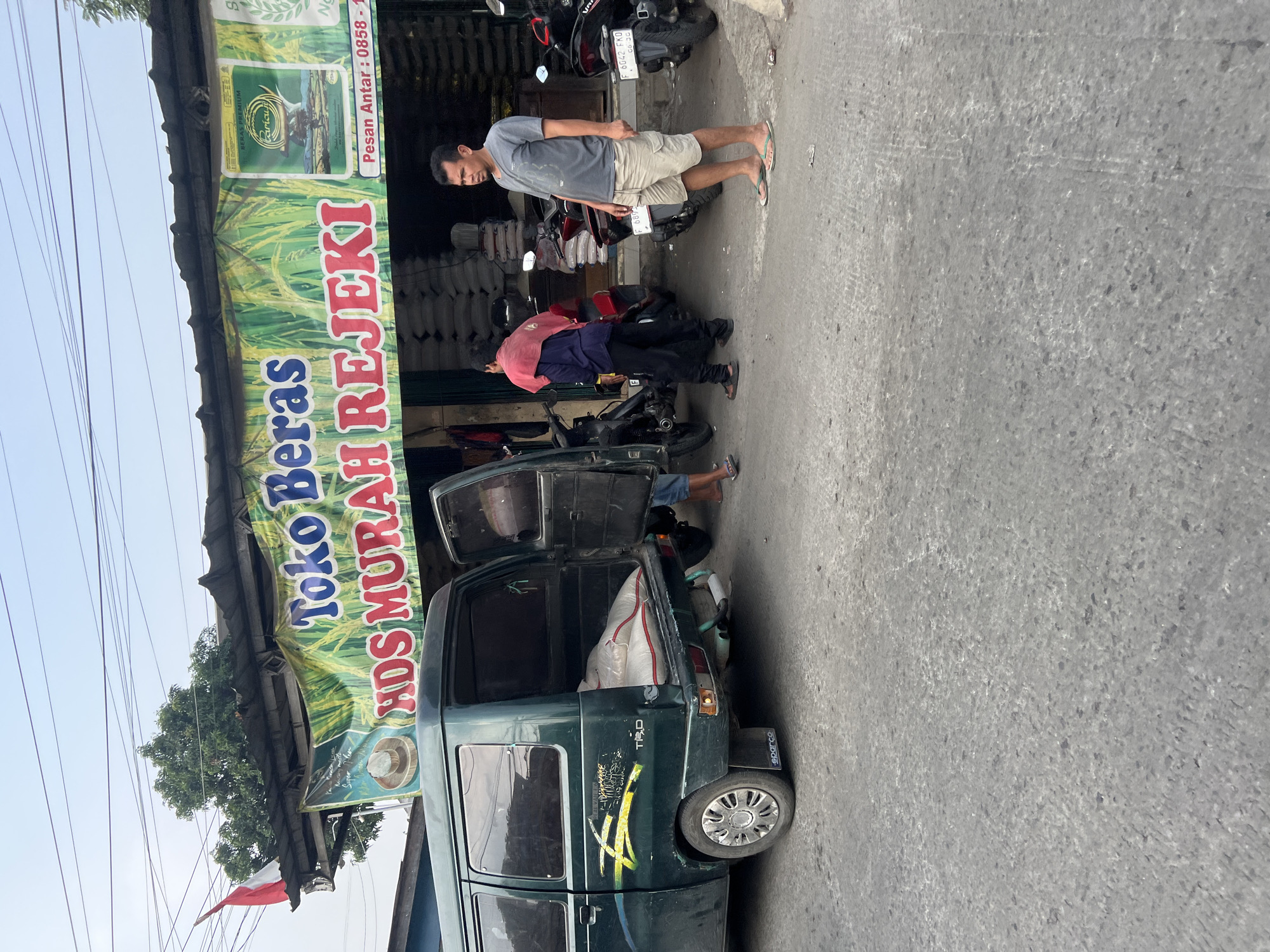 This image depicts a street scene in Citeureup, Indonesia, outside a rice shop. A green van is parked with its rear and side doors open, loaded with several large sacks that appear to be rice. Two men are near the van, one wearing a grey t-shirt and shorts, and another in a blue shirt and dark pants with a red cloth draped over his shoulders. Several motorcycles are parked in front of the shop. The shop itself has a banner overhead with Indonesian text that reads "Toko Beras HDS MURAH REJEKI" which translates to "HDS Cheap Fortune Rice Shop," and includes contact information for delivery. The street is paved, and the environment appears to be a commercial area with a moderate amount of activity.