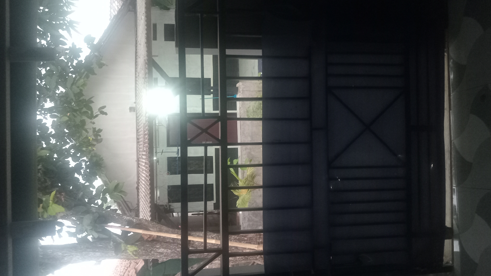 The image shows a view through a dark metal gate of a residential property. In the foreground, the gate is a prominent feature, composed of vertical bars and a more intricate lattice pattern at the bottom. Beyond the gate, a low concrete wall is visible, topped by a metal railing. Behind the railing, a building with a tiled roof is seen. The building has several windows with dark frames. A large tree with a textured trunk and leafy branches stands to the left of the building, casting shadows. Sunlight reflects brightly off the roof of the building and the surfaces of the windows, suggesting it is daytime. There are no people visible in the image. The setting appears to be a typical suburban or residential area, likely in Karawang, Indonesia, given the architectural style and vegetation.