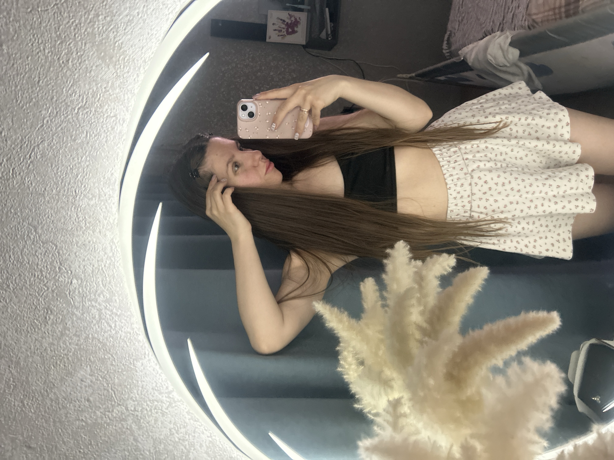 A young woman takes a selfie in a round, illuminated mirror. She wears a black crop top and a white floral mini skirt. Her long brown hair is parted down the middle. She holds a pink cell phone in her right hand. The background includes a bed, a table, and decorative pampas grass in front of the mirror. The image appears to be taken indoors, possibly in a bedroom. There is no information to indicate this image was taken in Klin, Russia.