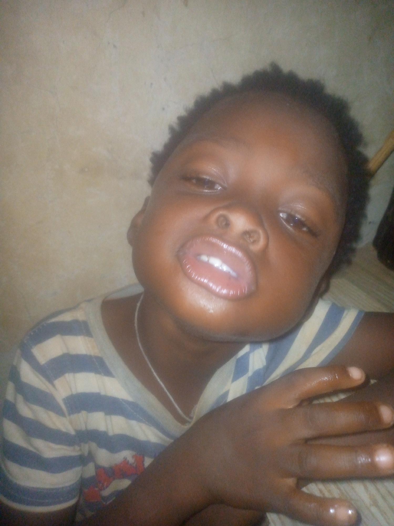 A medium, close-up shot of a young child with brown skin, short black hair, and full lips. The child wears a horizontally striped, light blue and white shirt and a thin, silver necklace. The child’s right hand rests in front of them. The background appears to be a mottled off-white wall with a wooden element visible on the right side. The location appears to be indoors in an unknown city.