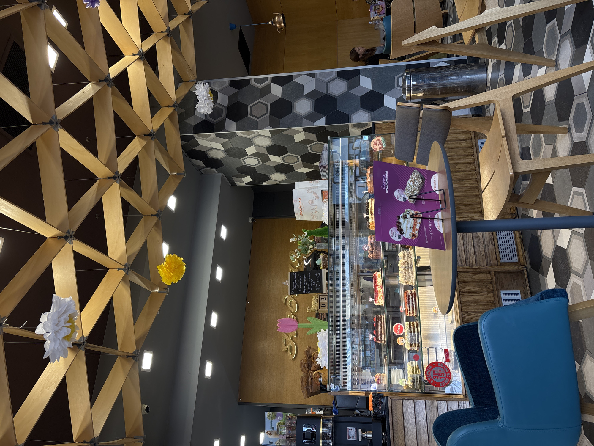 The interior of a cafe in Varna, Bulgaria is shown with a hexagonal patterned tile floor. There are wooden tables and chairs. There is a display case full of pastries, with advertisements for the pastries visible. A wooden framework is on the ceiling with flowers. In the background, a person is sitting at a table.