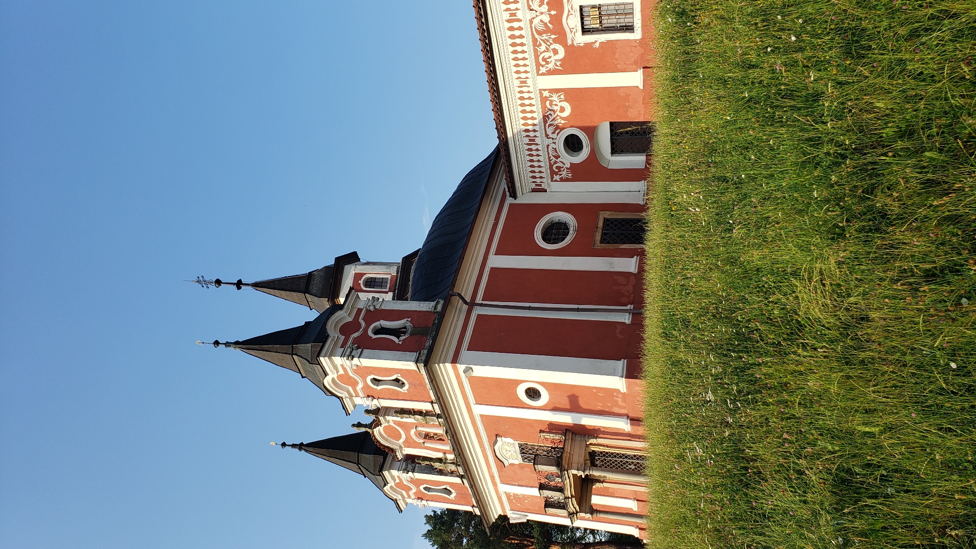 This is a medium shot, eye-level, vertical, natural light outdoor photograph of the Church of the Nativity of the Virgin Mary in Prešov, Slovakia. The church is painted red with white trim and features multiple spires and ornate architectural details. The building is partially visible, with a large section of lush green grass and wildflowers in the foreground. The sky is a clear, bright blue. The photograph captures the church from a side angle, emphasizing its colorful facade and towering spires. There are no people or animals visible in the image.