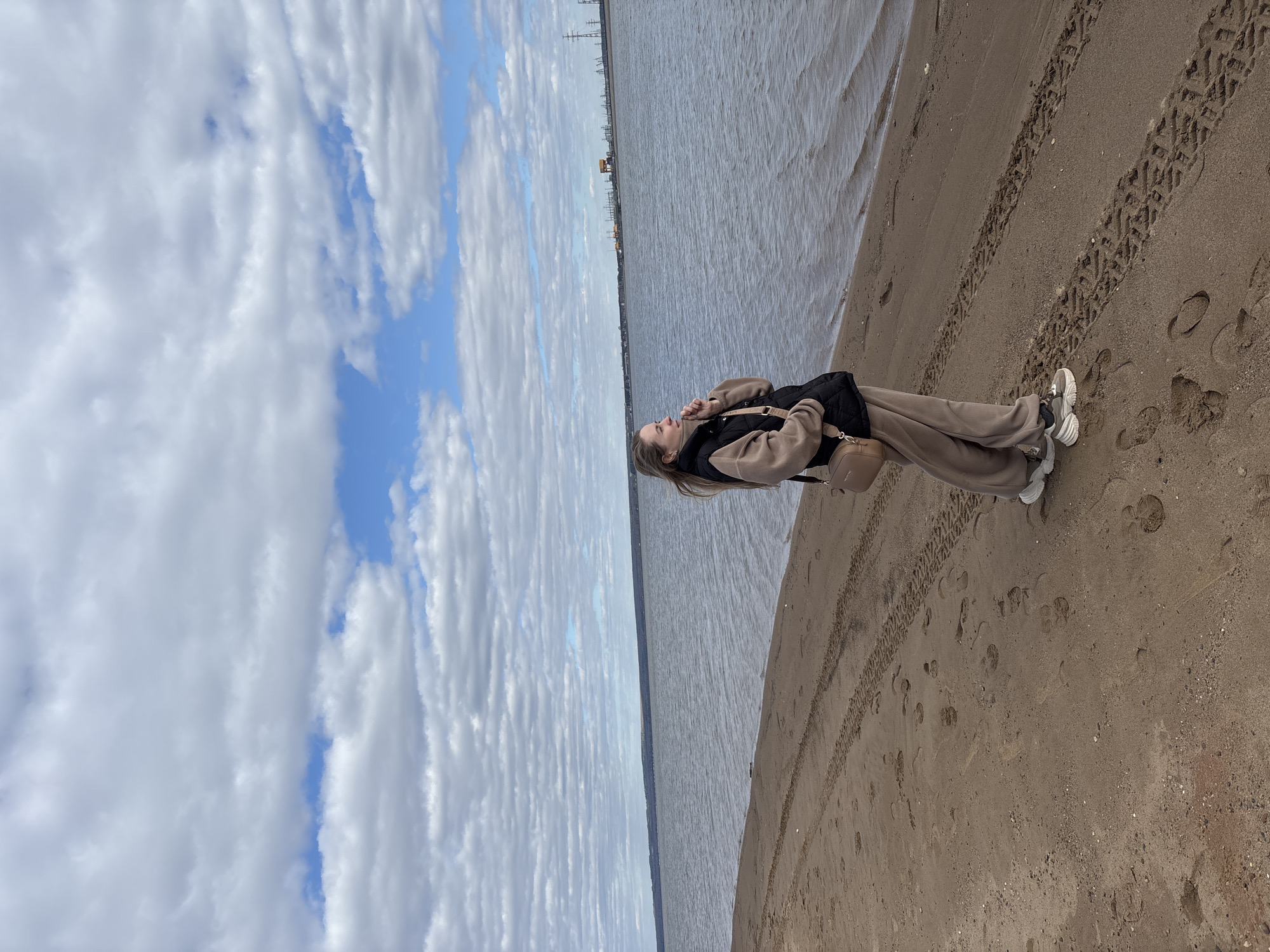 A young woman stands on a sandy beach with calm water to her right and a partly cloudy sky to her left. The woman is wearing light brown trousers, a matching hooded jacket, a black vest, and white sneakers. She is holding a small brown bag. Tire tracks and footprints are visible in the sand. In the distance, across the water, are industrial structures and power lines, suggesting an urban setting. The city is Noviy, Russia.