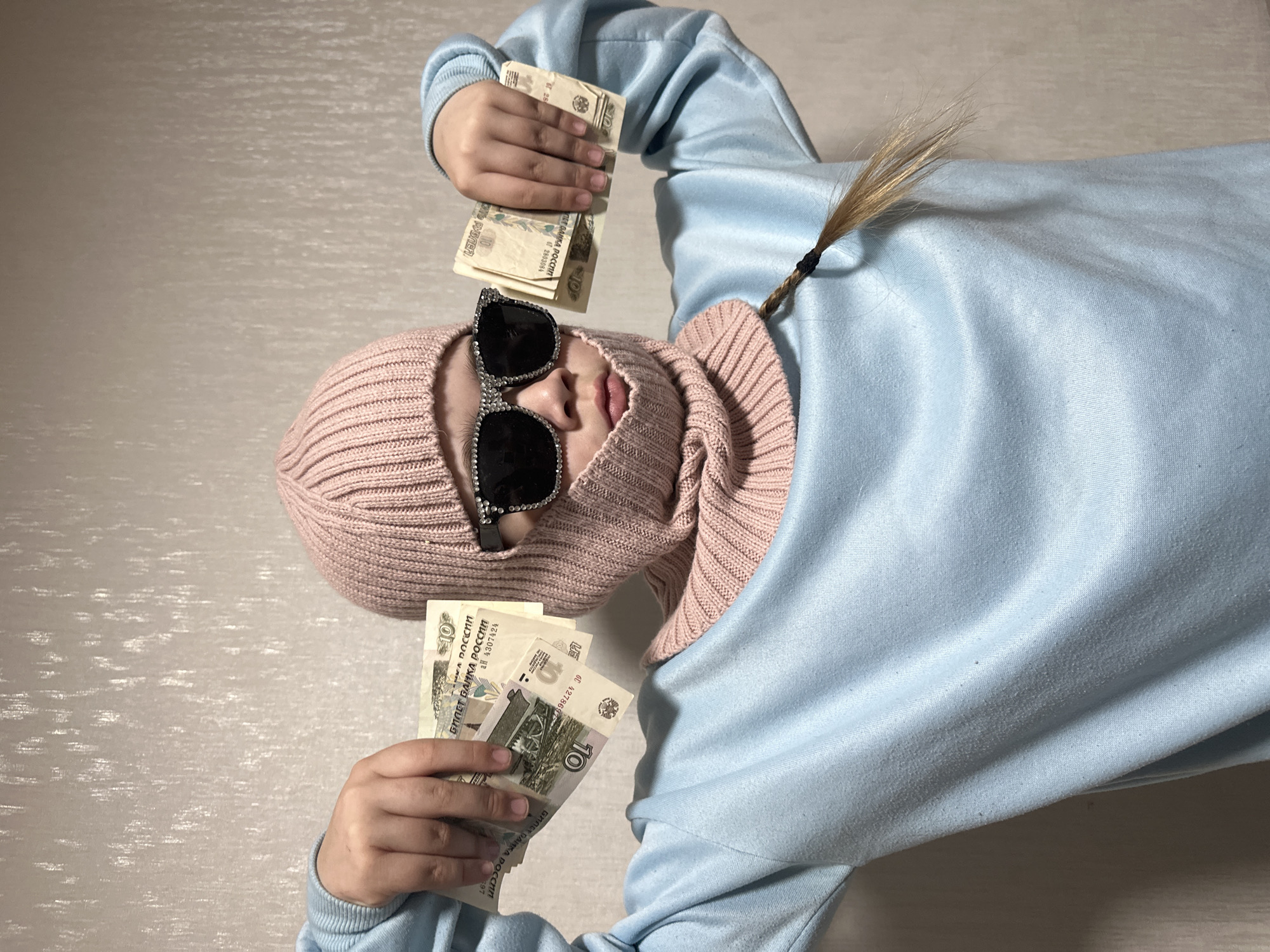 A person is depicted lying on their back on a light-colored, textured surface, possibly a bed or floor. They are wearing a pink knitted balaclava that covers their head and neck, with an opening revealing their nose and mouth. Large black sunglasses adorned with sparkling embellishments cover their eyes. The person is dressed in a relaxed-fit, light blue long-sleeved top or hoodie.
In both hands, the individual is holding and displaying stacks of Russian 10 Ruble banknotes. A small ponytail of light brown or blonde hair, secured with a black elastic band, is visible emerging from the back of the balaclava. The posture and display of money suggest a confident, playful, or boastful attitude.
The banknotes clearly show the text "БИЛЕТ БАНКА РОССИИ" (Bank of Russia Ticket), the denomination "10", and "РУБЛЕЙ" (Rubles). Specific serial numbers and codes such as "АН 2803084", "БН 4278697", and "АН 4307424" are visible on different notes. The scene is set indoors, with the light, neutral background providing a simple context. The currency confirms the location context of Chaikovsky, Russia.