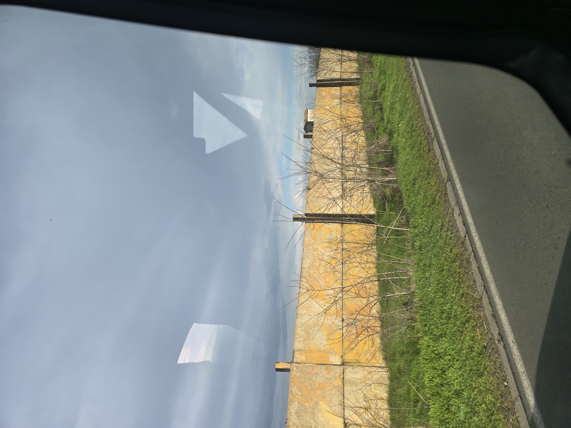 The image, taken from within a vehicle, shows a section of a road in Kabile, Bulgaria. Along the roadside is a weathered, yellow-painted concrete wall or barrier, possibly a property boundary or retaining wall. Sparse, leafless trees or shrubs grow along the base of the wall. Next to the wall is a strip of green vegetation. The road surface is asphalt, with a light-colored curb or edge marking its boundary. The sky is overcast with light blue and gray. There are reflections of the sky and possibly interior vehicle components on the window through which the photograph was taken.