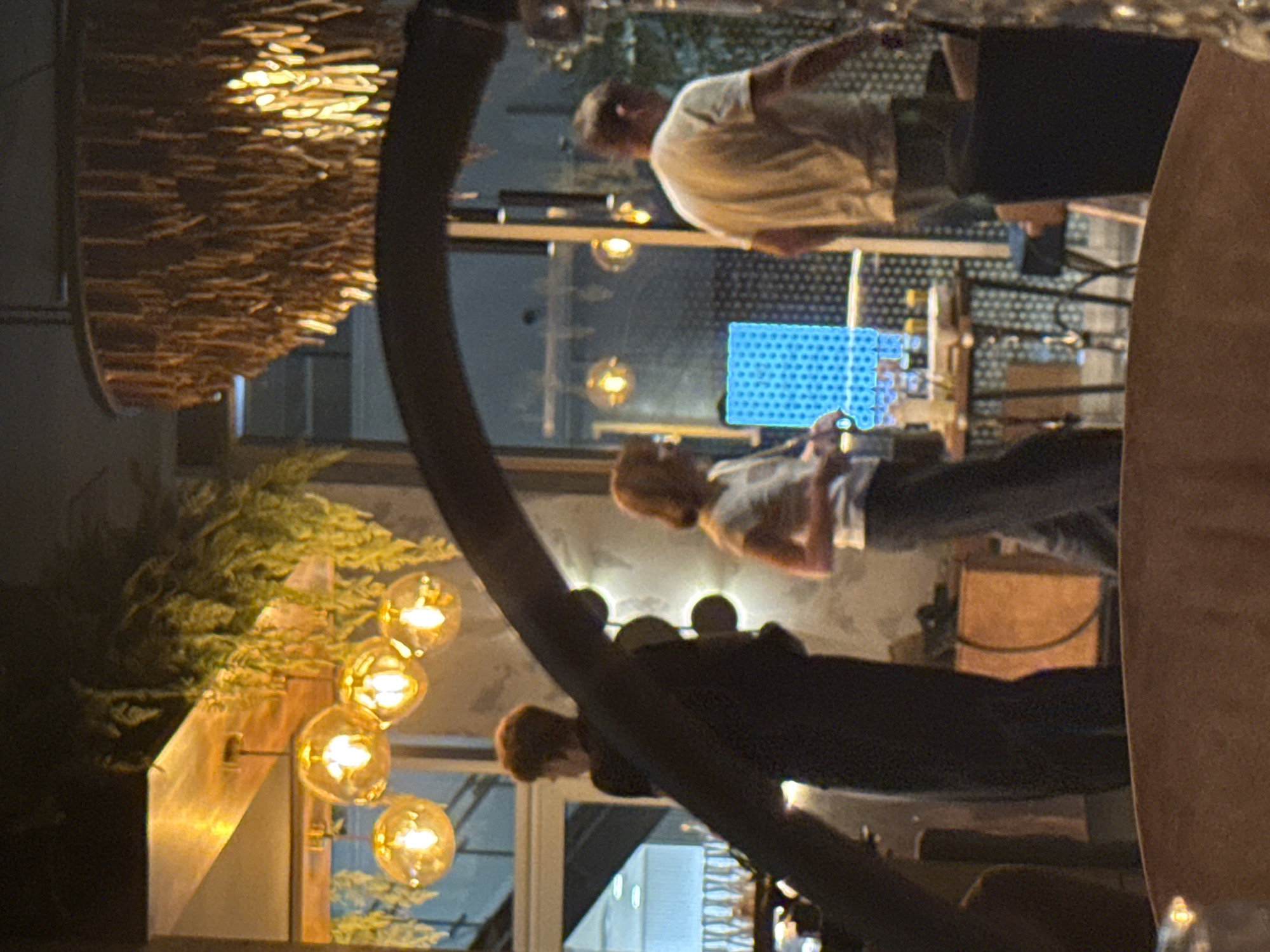 The image depicts an indoor scene, likely a restaurant or bar, with ambient lighting from hanging fixtures. In the foreground, a dark, curved object partially obscures the view. Several people are visible in the midground, including a person in a dark outfit on the left, a woman in a light-colored shirt and dark pants in the center, and a person in a light-colored t-shirt on the right, facing away from the camera. Behind them, there are tables, some with drinks and a blue, illuminated geometric display. Hanging light fixtures with spherical shades are present in the upper portion of the image, along with greenery. The scene suggests a social gathering in a dimly lit, contemporary setting. There is no information available in the image to identify the specific location in Bratislava, Slovakia.