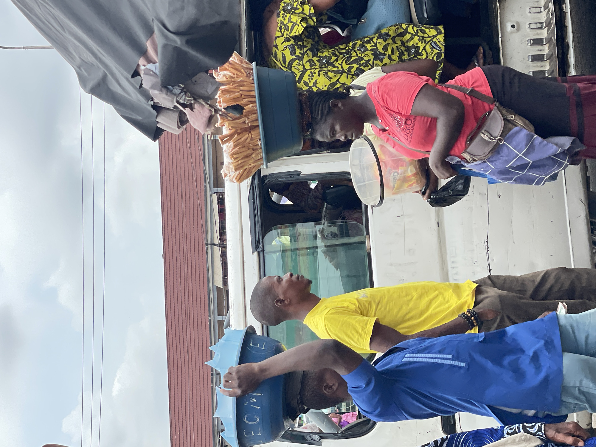 A group of people are gathered around a white vehicle loaded with goods in Onitsha, Nigeria. Two men stand in the foreground, one wearing a yellow shirt and the other in blue, both carrying blue containers on their heads. A woman in a pink shirt stands in the truck bed, along with another person in a yellow patterned fabric. The truck appears to be packed with various items, including long, thin, beige objects. The environment includes a building with a reddish-brown roof, a cloudy sky, and what appears to be an overhead cloth.