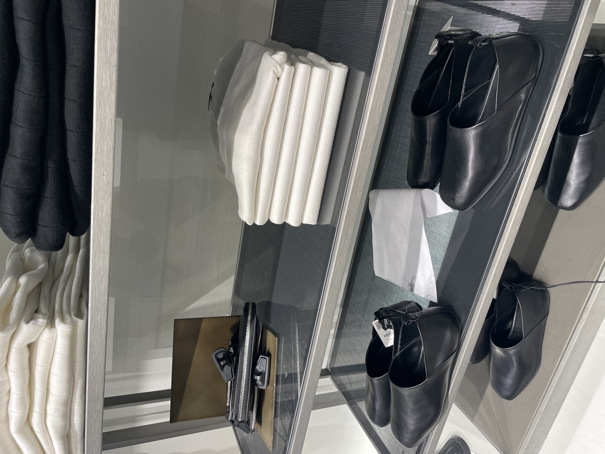 This image showcases a display of clothing and accessories, likely in a retail setting. On the left, stacked garments in black and white are visible. To the right, neatly folded white textiles are positioned on a shelf. Further right, a selection of black leather mules are displayed, some bearing tags. A small, rectangular brown bag with black zippers is also present. The scene is well-lit, suggesting an indoor environment, possibly a boutique or department store.