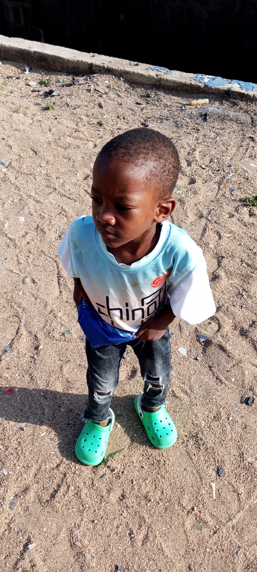 A young Black boy stands on a sandy ground in what appears to be an outdoor urban environment in Jalingo, Nigeria. He is wearing a light blue t-shirt with white sleeves and a graphic print, black ripped jeans, and bright green Crocs. His left hand is inside his pants pocket. The background shows a concrete barrier or wall with some debris. The lighting suggests it is daytime.
