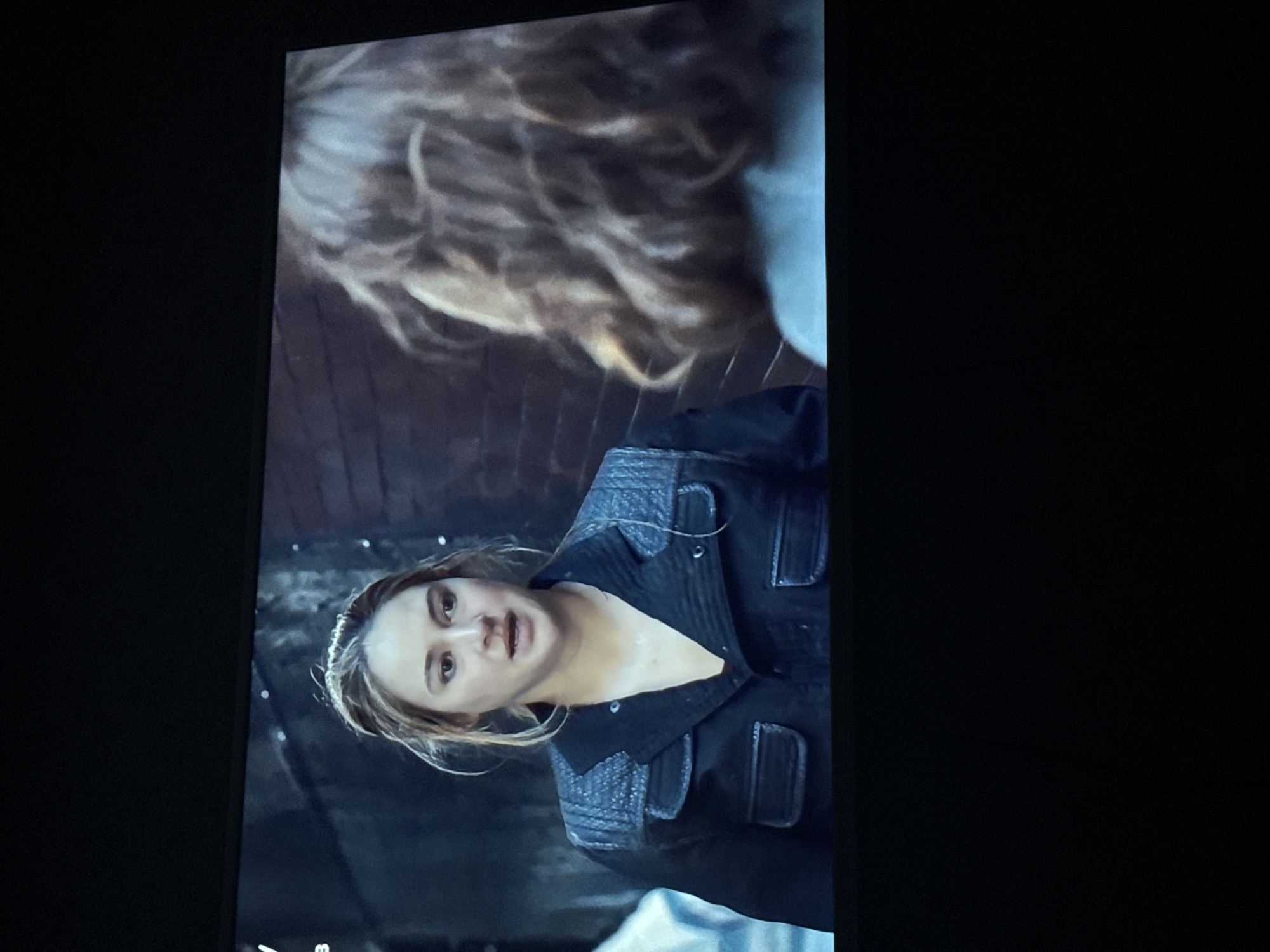 The image shows a scene from a film or television show, featuring a woman with light brown hair and fair skin, wearing a dark-colored jacket with a button-down front. The jacket has textured shoulder and pocket details. She is looking slightly upward with an expression of concern or distress. Above her is a person with blond hair, appearing out of focus in the background. The environment is dimly lit, suggesting an indoor setting with a brick or textured wall. The lighting is stark, creating shadows and emphasizing the woman's facial expression. There is no information provided to suggest this image was taken in the city of Mytishchi, Russia.