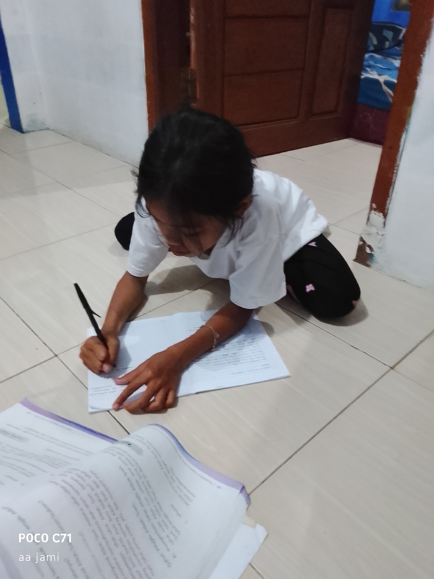 In a residential setting, likely a home in Tasikmalaya, Indonesia, a young girl is seen on the floor, engaged in writing. She is wearing a white t-shirt and black pants adorned with small pink heart designs. The girl is kneeling and leaning forward, her focus directed at a stack of papers spread out before her. In her right hand, she holds a black pen, actively writing on the top sheet of paper. Her left hand rests on the papers, stabilizing them as she works. The floor appears to be tiled in a light beige color. In the background, a wooden door is visible, and to the right, there is a glimpse of bedding with blue and red patterns. In the foreground, another open book lies on the floor, with text visible on its pages. The lighting suggests an indoor environment, possibly during the day.