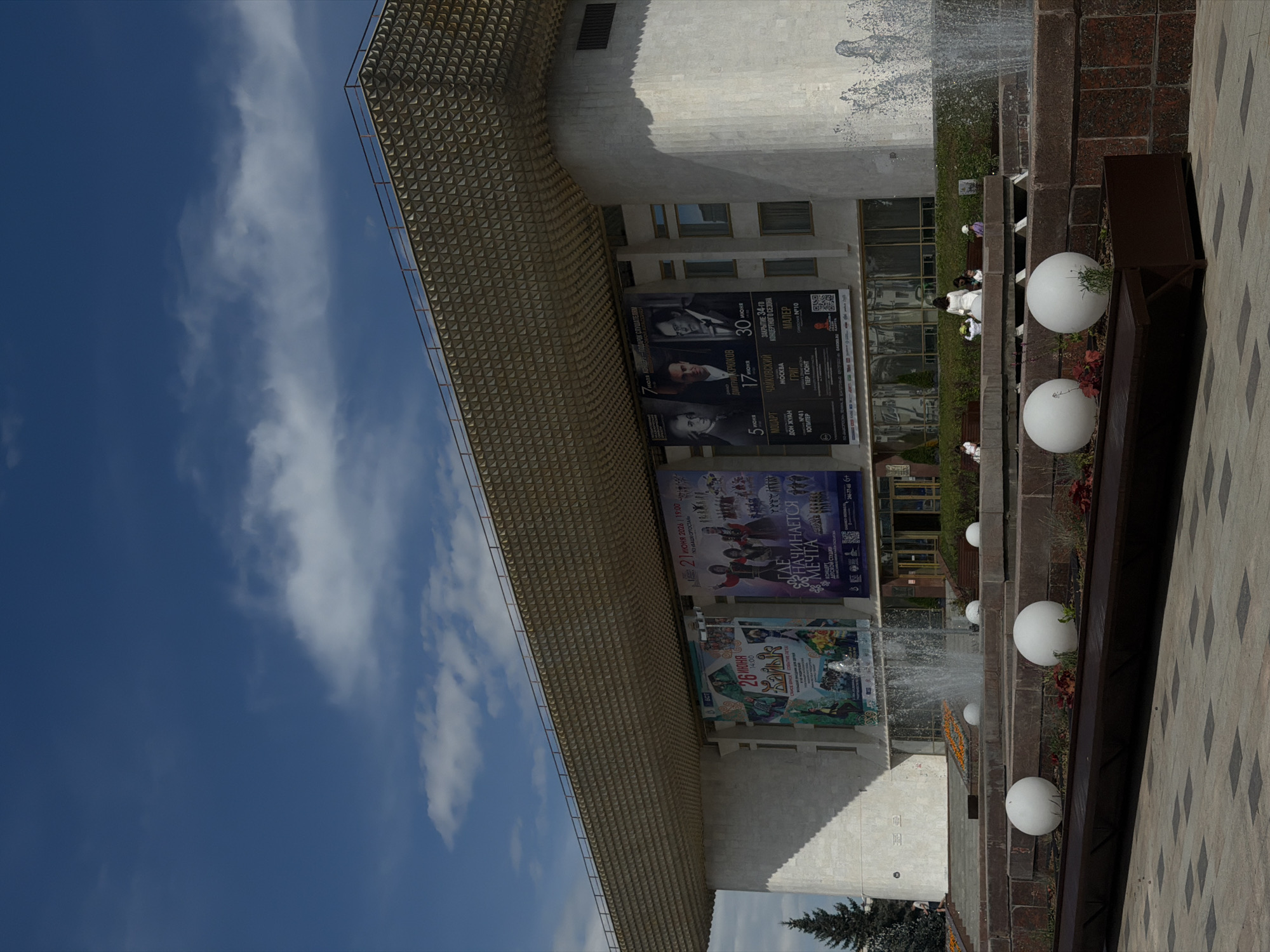This photograph captures an outdoor scene in Ufa, Russia, on a bright, sunny day with scattered white clouds against a deep blue sky. The primary focus is the facade of a modern building, possibly a cultural center or theater, with a distinctive patterned overhang. Below this overhang, several large posters are displayed, advertising upcoming performances.
The posters detail events featuring classical music, with names like Mozart, Tchaikovsky, and Grig appearing. Specific dates and times are visible, such as "26 июня 14:00" and "21 ИЮНЯ 2026 19:00," indicating the content is for June events. Some posters also include images of performers, suggesting classical concerts or opera.
In the foreground and to the right, a tiered fountain area is visible. Water is spraying from the fountain, creating a dynamic element in the scene. Large, white spherical decorations, possibly planters or art installations, are placed on ledges along the fountain. Several people are seated on benches in the background, observing the fountain or waiting. One person in a white dress is standing near the fountain. The overall atmosphere appears to be calm and pleasant, suggesting a public space utilized for leisure and cultural engagement. The tiled pavement in the foreground adds to the urban setting.