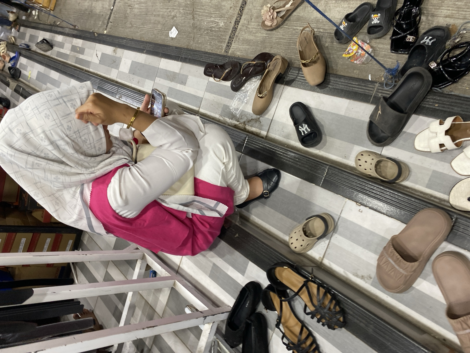 In Lamteumen Barat, Indonesia, a person wearing a white hijab and a pink and white outfit is crouching on a tiled floor, looking at a smartphone. Various footwear, including sandals, flip-flops, and clogs, are scattered around the immediate area on the floor. The background shows a striped tiled floor and metal railings, suggesting a commercial or market setting.