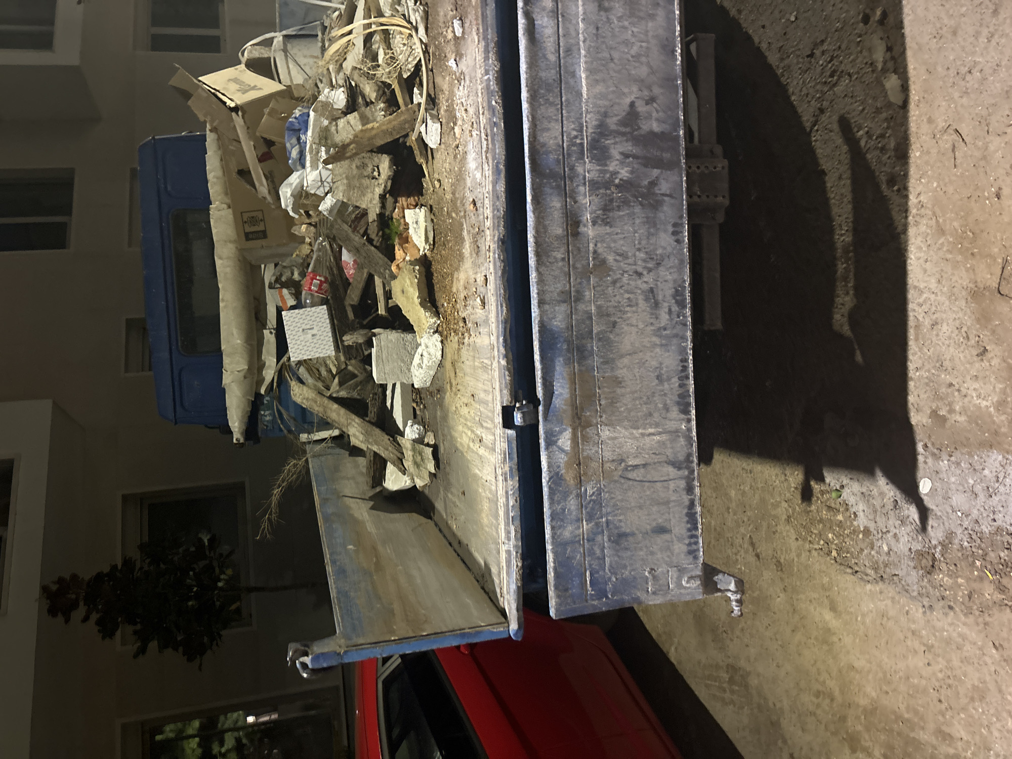In Budva, Montenegro, a blue flatbed truck is parked on a paved surface, likely a street or parking lot, at night. The truck's bed is filled with construction debris, including broken concrete, wood fragments, and cardboard boxes. A red car is parked to the left of the truck, partially visible. A small tree stands in front of a building in the background, which appears to be an apartment complex with several windows. The lighting suggests artificial light sources are illuminating the scene.